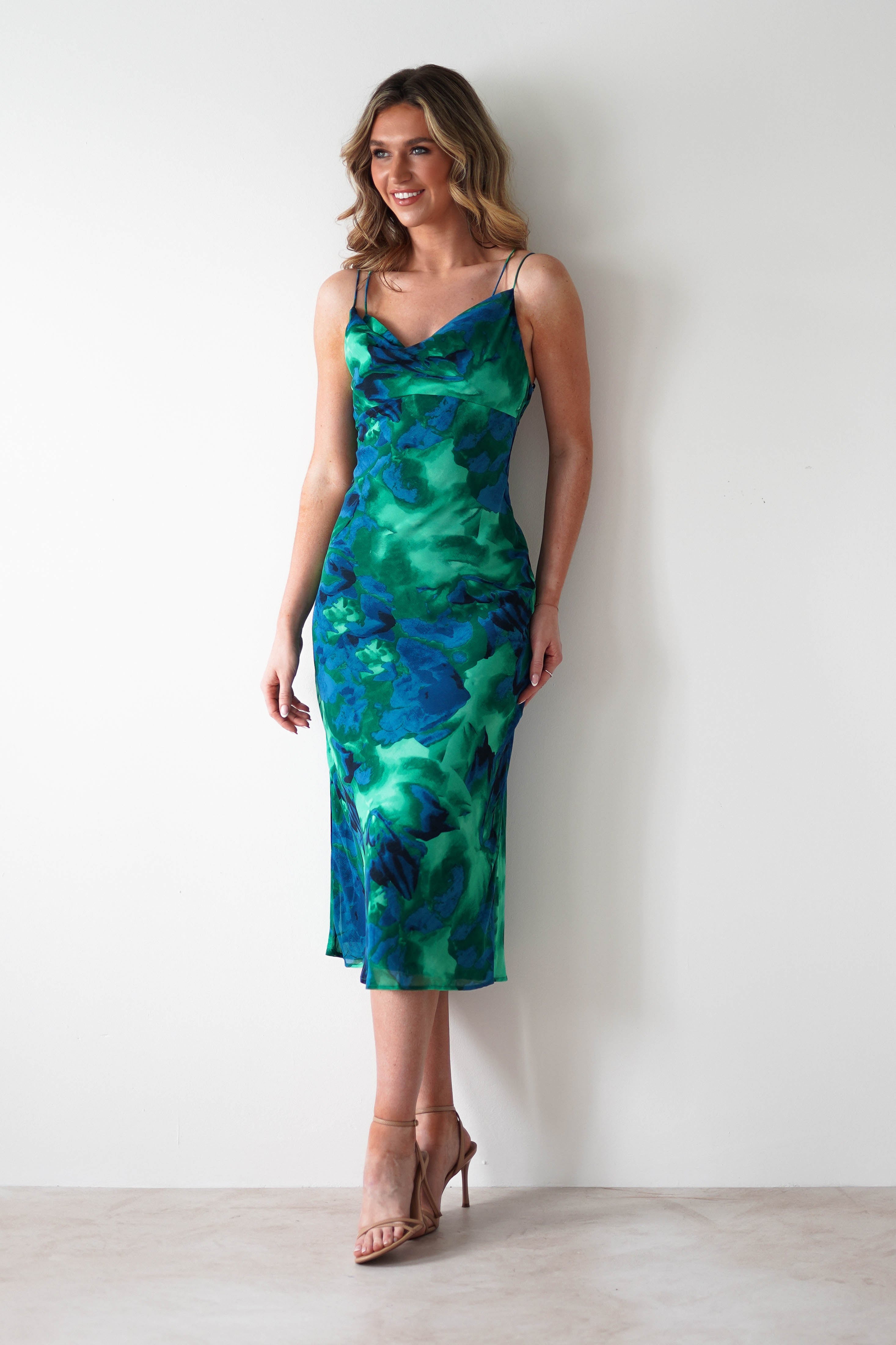 A woman in a printed midi chiffon wedding guest dress with thin straps stands against a white wall, smiling and wearing nude high-heeled sandals.