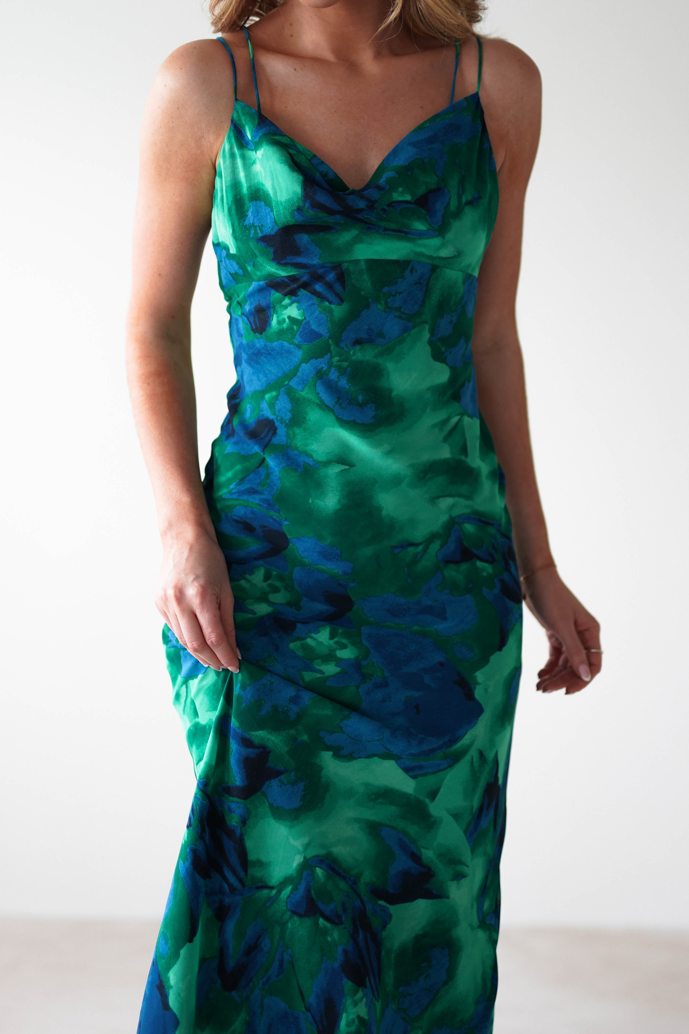 A woman wearing a sleeveless, green and blue abstract-patterned satin dress with thin straps, perfect for occasions like a wedding guest dress, standing against a plain white background.