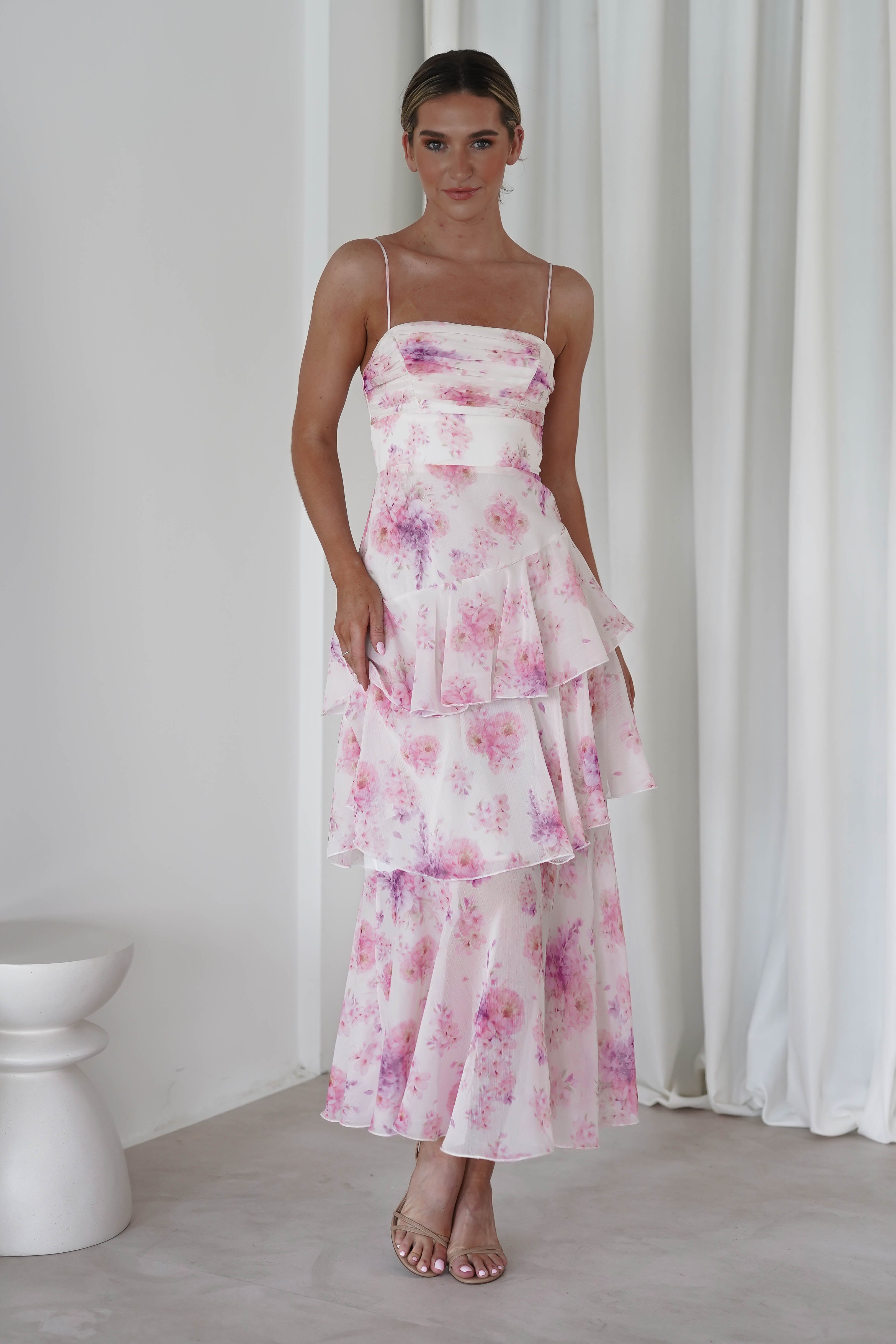 In a softly lit room, someone elegantly poses in a sleeveless, tiered pink floral midi dress with enchanting purple patterns. One hand rests on their hip, embodying grace. The backdrop of white curtains complements this perfect wedding guest dress look.