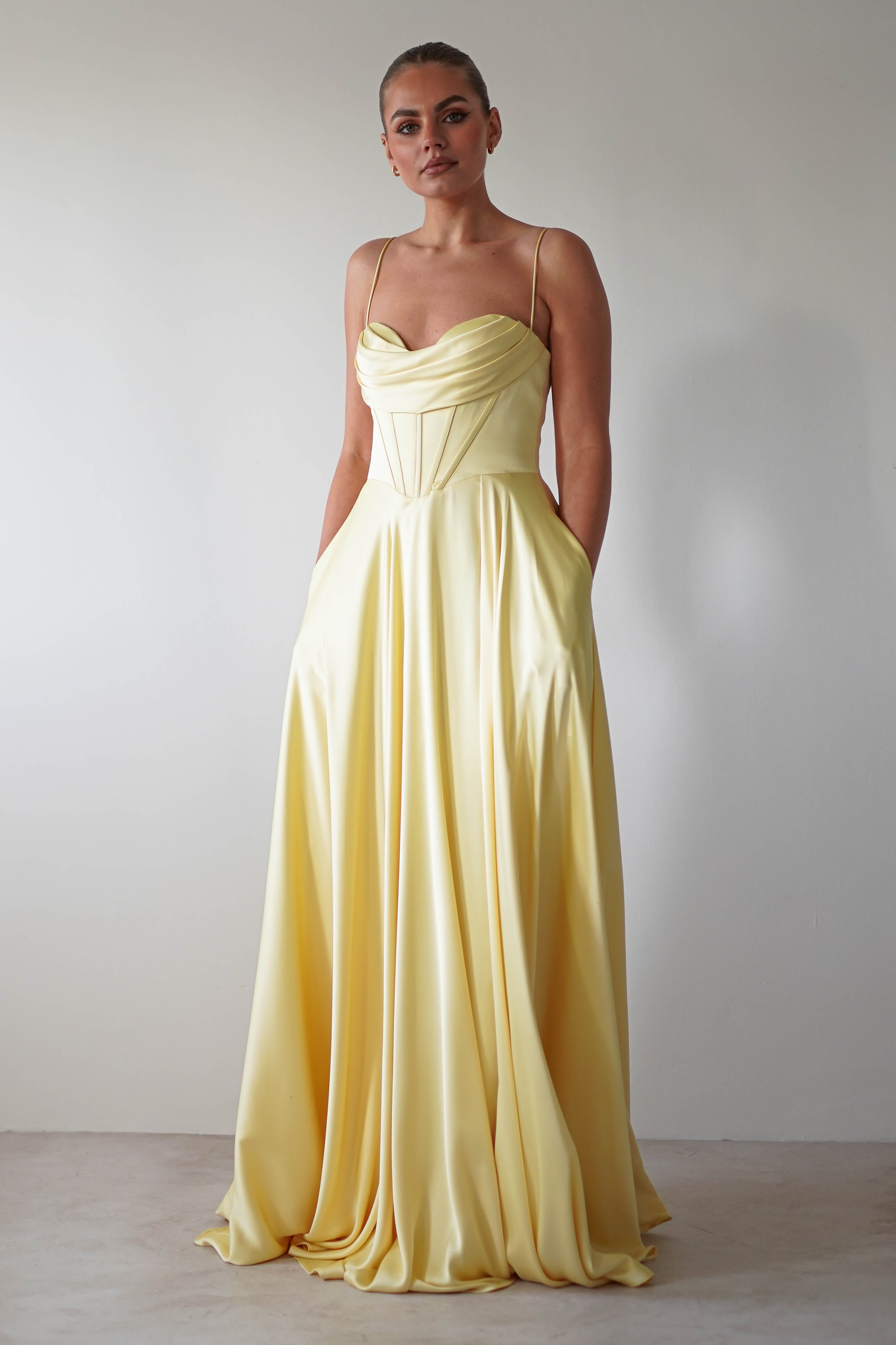 A person stands against a white background, wearing a flowing yellow satin gown with thin straps and corset detailing on the draped bodice. The dress has a floor-length skirt and their hands rest in the dress pockets. The lighting is soft and evenly distributed.