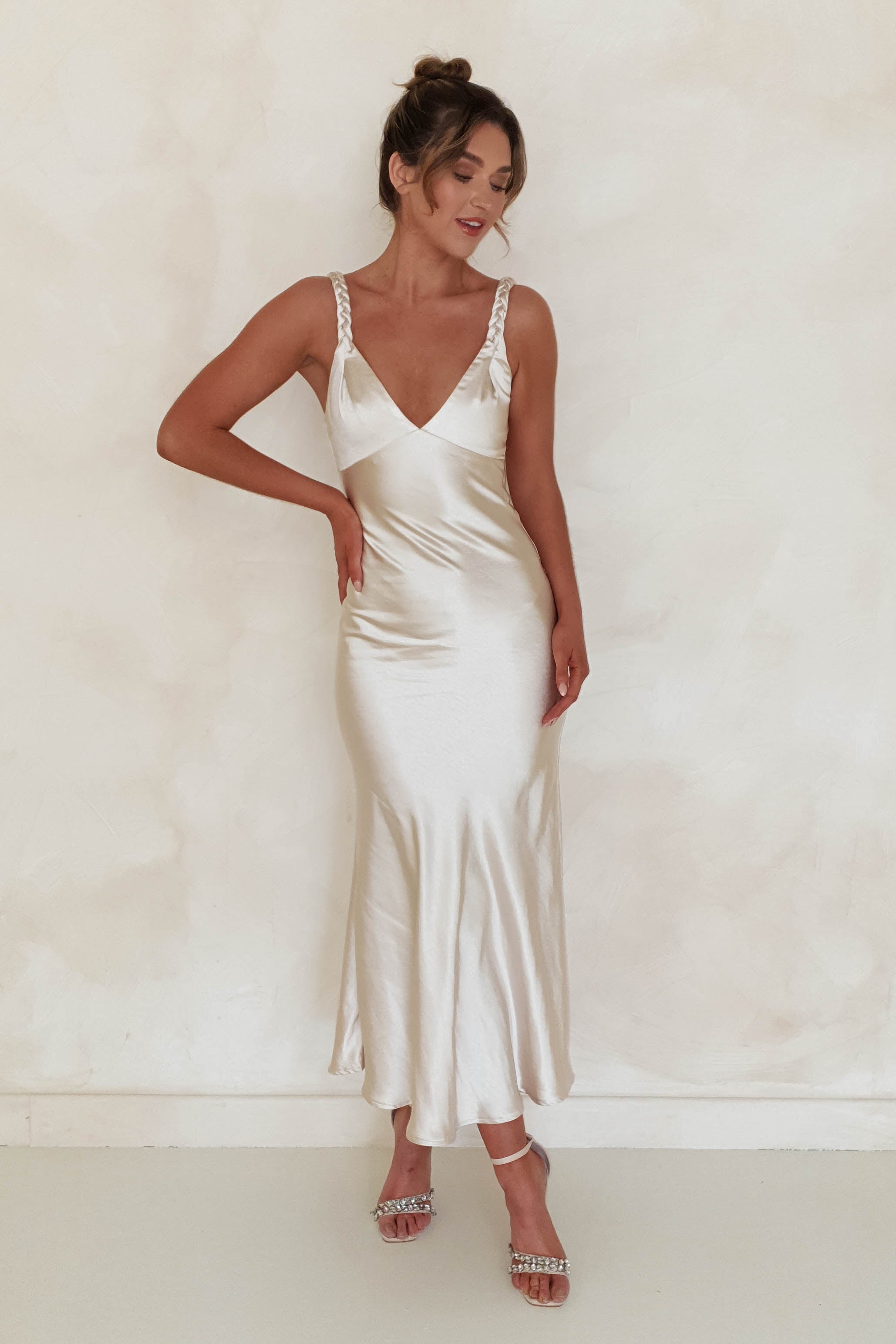 A woman poses against a neutral background, showcasing a sleek, cream-colored satin maxi dress with plaited straps. Her hair is styled in an updo, and she stands confidently in heeled sandals, exuding elegance perfect for special occasions.