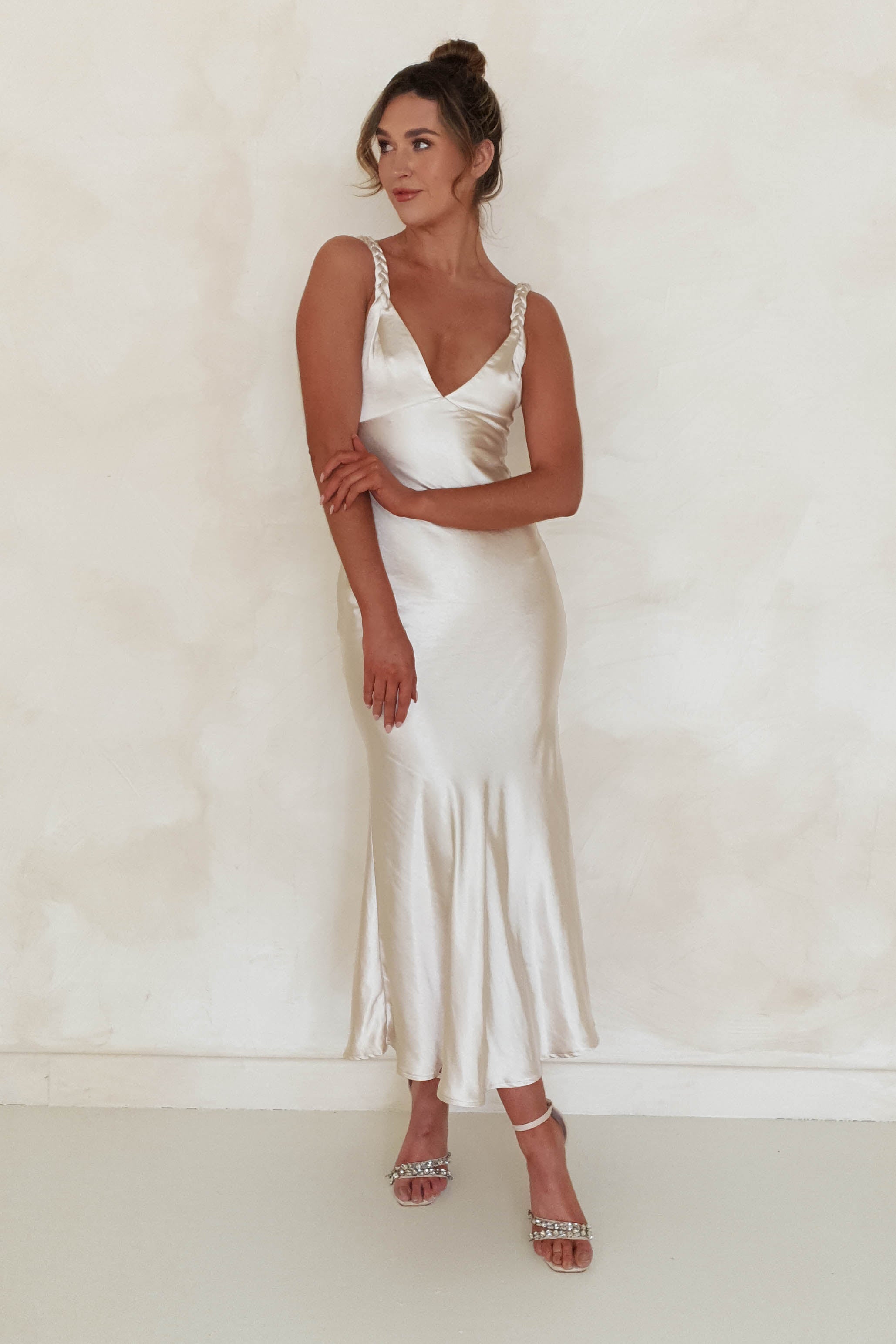 A person stands against a light background wearing a sleeveless, satin maxi dress featuring plaited straps. Their hair is up as they gaze to the side, one arm crossed and the other resting on it. Strappy heels complete the ensemble, perfect for special occasions.