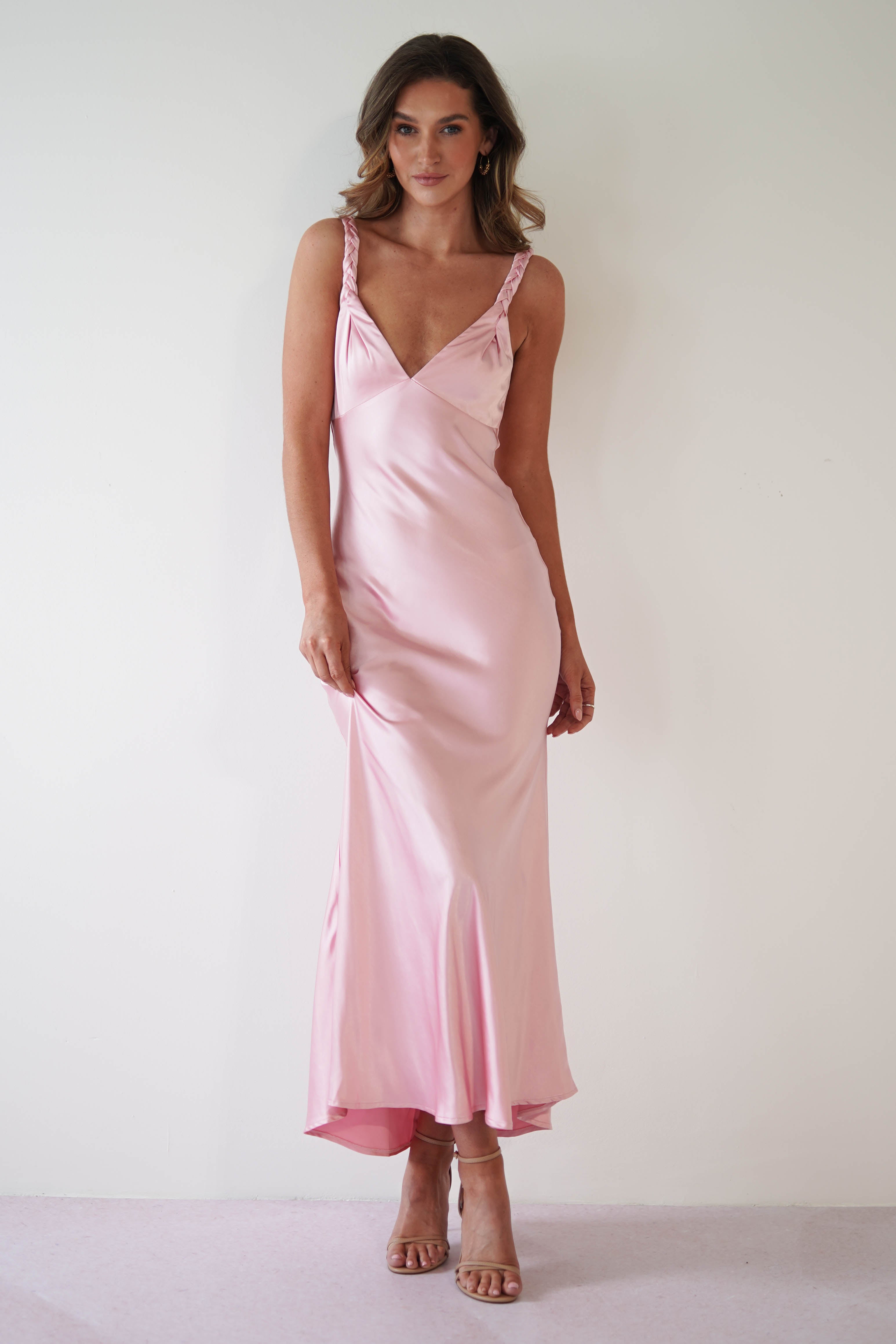 Woman in a long, satin maxi dress poses against a plain white background. The silky pink gown, perfect as a wedding guest dress, features thin straps and a flowing design. She stands confidently, looking towards the camera with minimal accessories and nude heels.