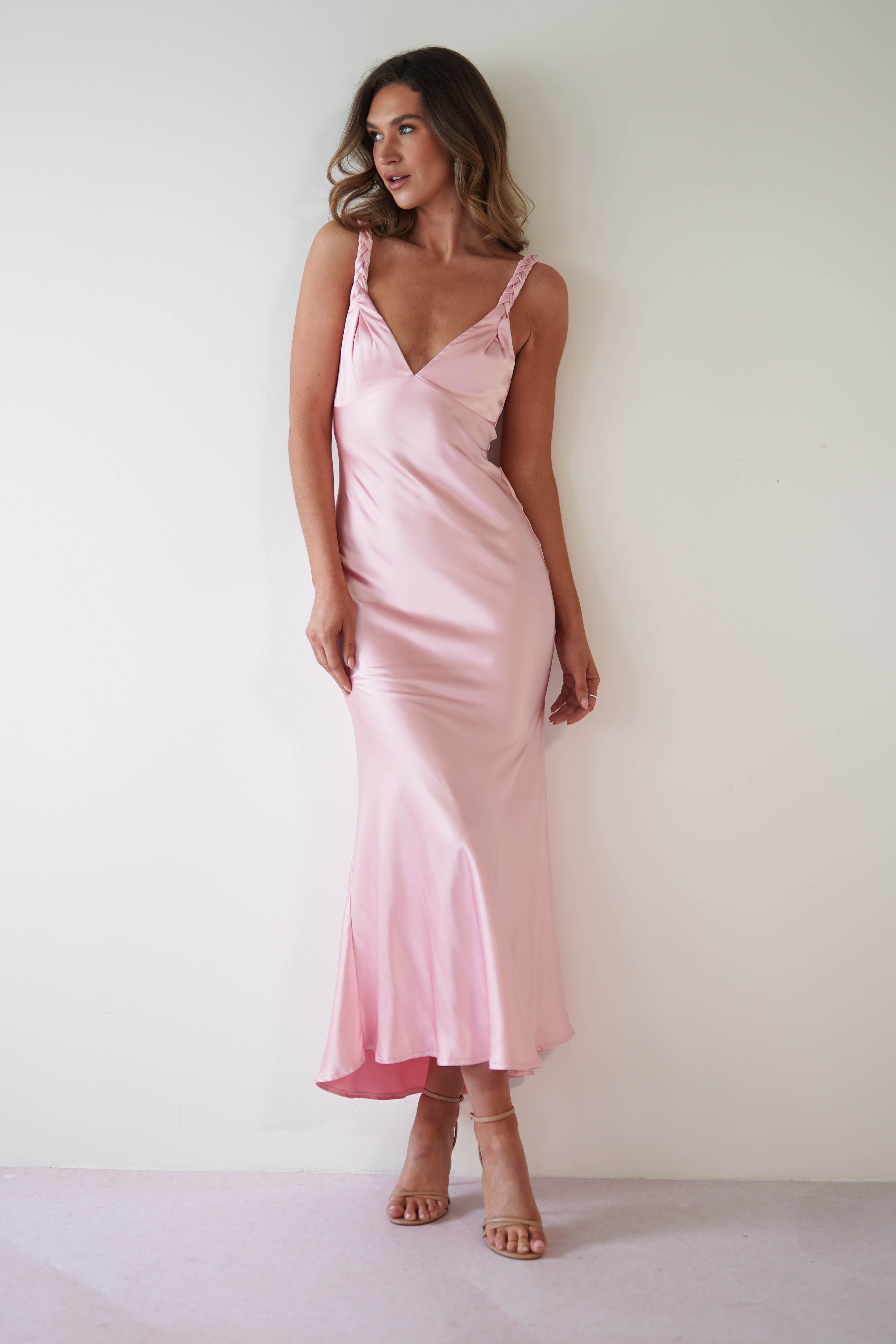 A woman in a pink satin maxi dress stands against a plain white background. She has long, wavy hair and is wearing nude heels. The sleeveless dress features a V-neckline and falls to her ankles, perfect for a wedding guest ensemble. She looks elegantly to her right.