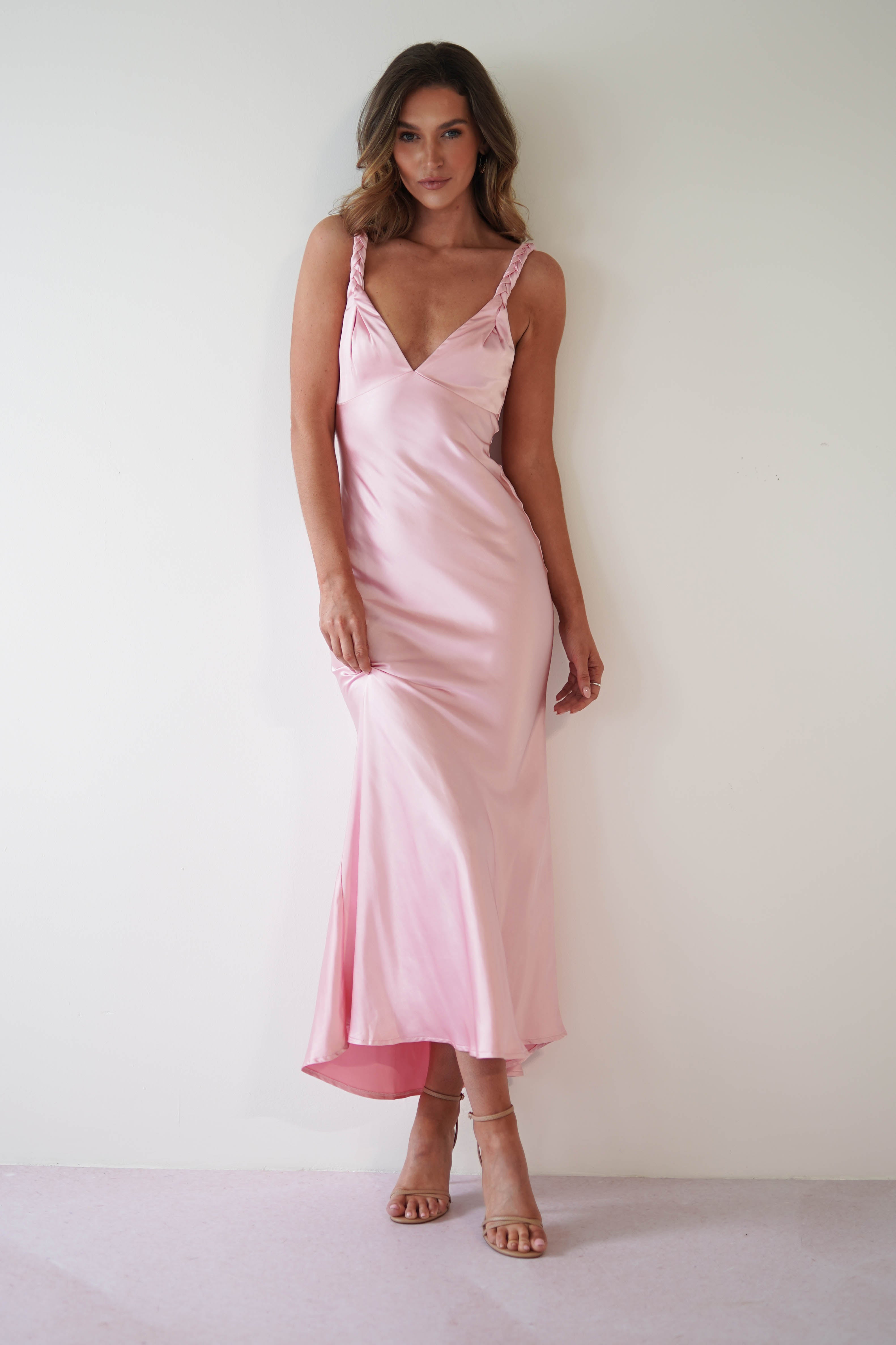 A woman stands against a white wall in a pink satin maxi dress and nude heels. She gazes at the camera with a gentle smile, one hand slightly lifting the dress at her side. Perfect as a bridesmaids dress, her wavy hair elegantly falls over her shoulders.