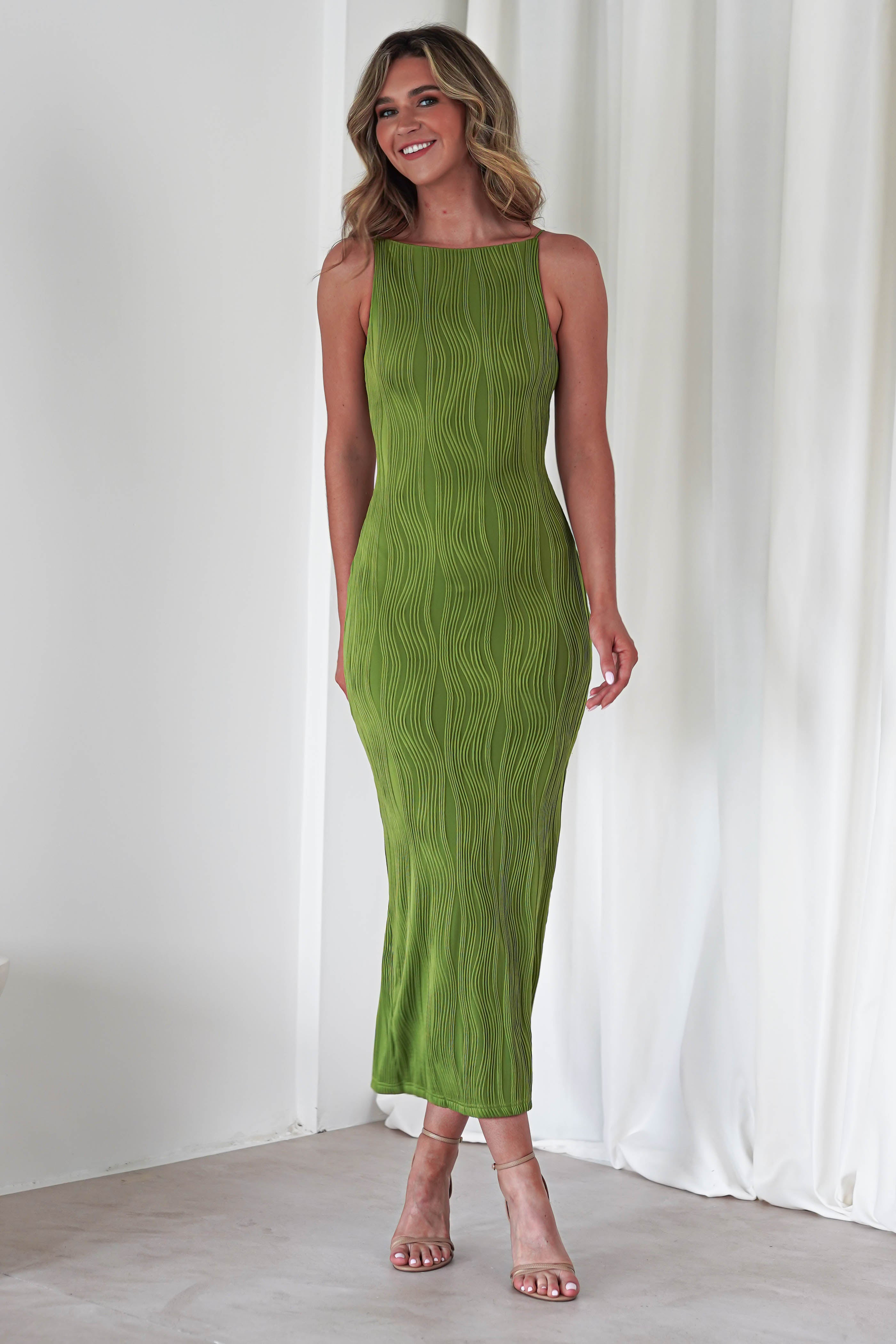 A woman stands indoors in a green, wavy textured midaxi dress, perfect for a wedding guest. The sleeveless dress pairs with strappy nude heels. She smiles with her wavy blonde hair, white curtains in the background.