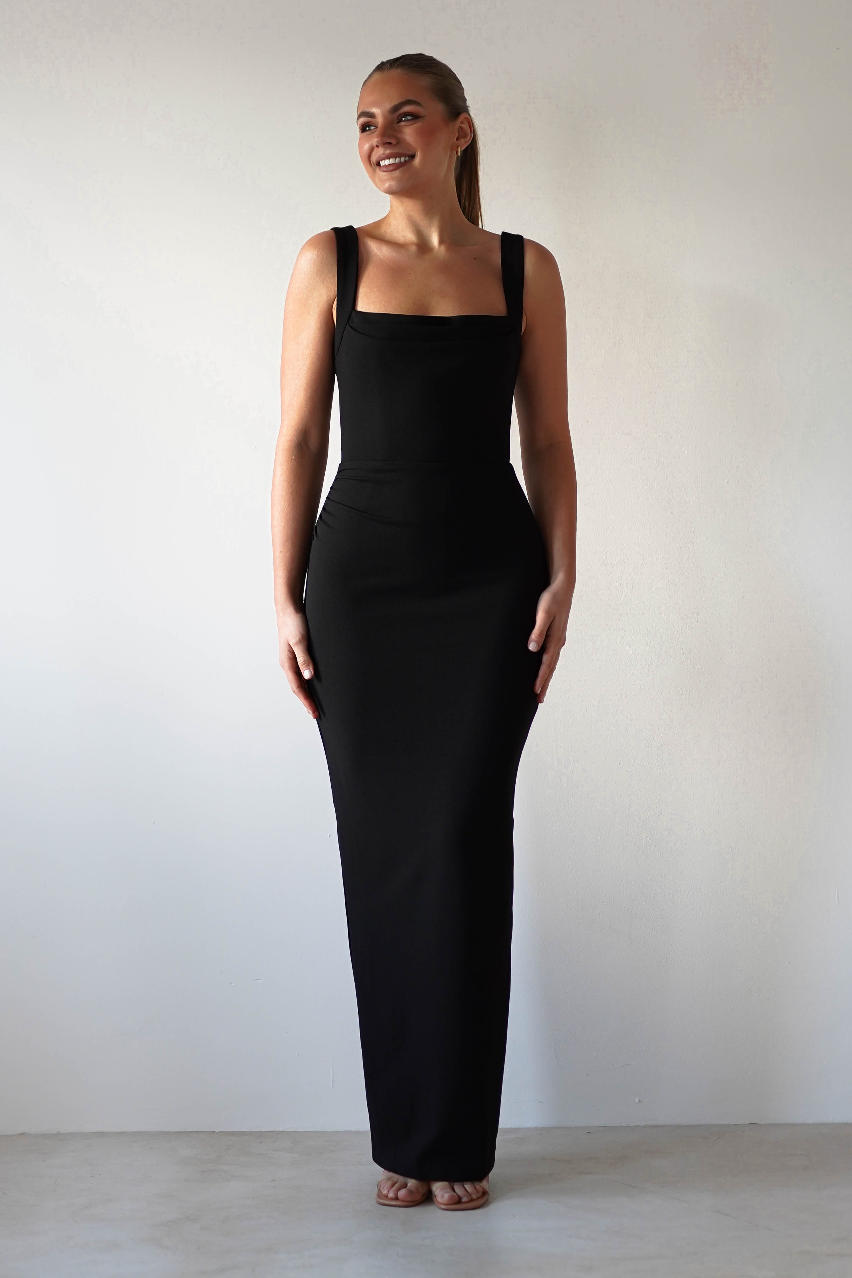 A person stands against a plain background wearing a glamorous, floor-length black dress with wide straps. The bodycon fit hugs their form elegantly. Their long hair is pulled back, and they’re smiling with hands resting by their sides.