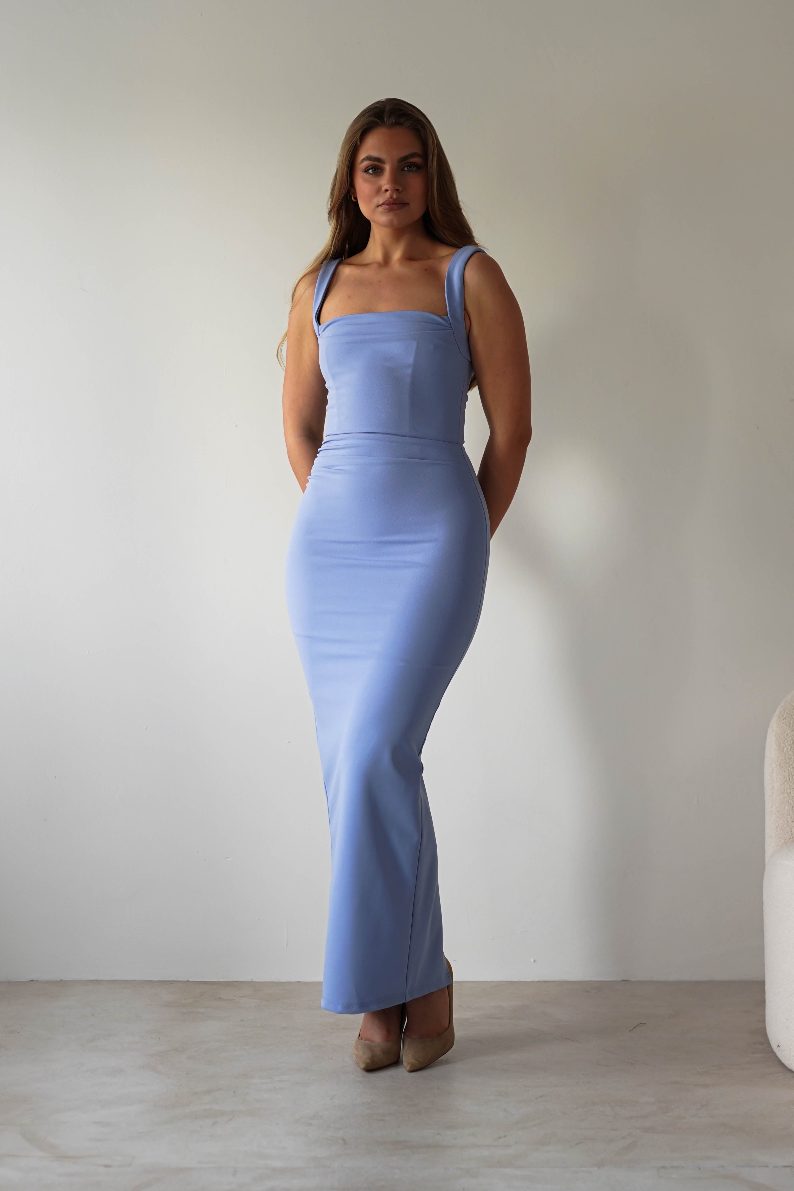 A woman stands indoors against a plain white wall, wearing a light blue square neck bodycon dress and beige heels. Her arms are behind her back as she looks toward the camera—a stylish look perfect for a wedding guest dress.