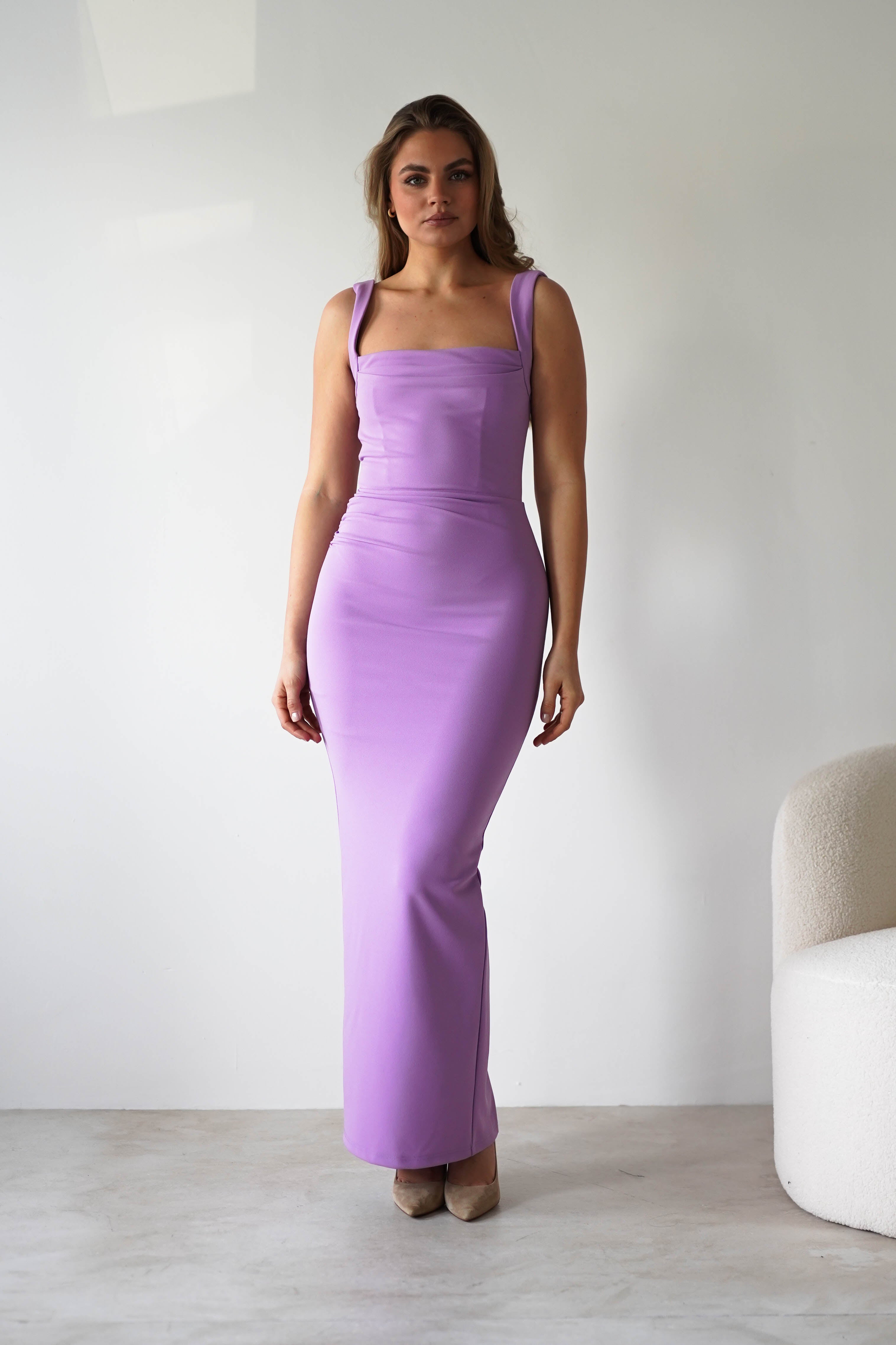 A woman stands indoors against a plain white wall, wearing a lilac square neck dress with a flattering bodycon fit and beige heels. She has loose, wavy hair and a neutral expression. A white, textured chair is partially visible on the right.