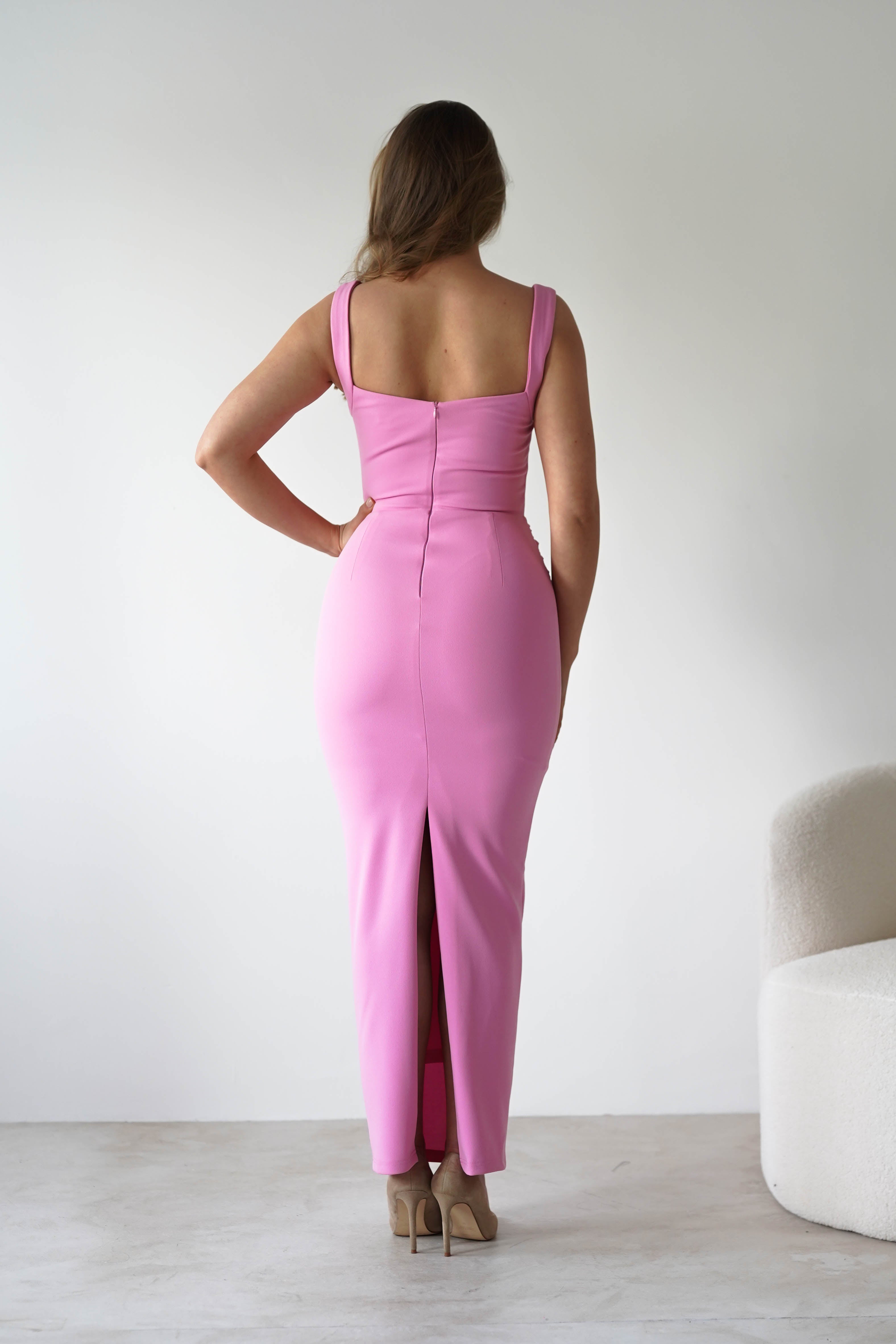 A woman wearing a fitted, sleeveless pink square neck dress stands facing away from the camera in a minimalistic room with white walls and a white chair—a chic choice for a wedding guest.