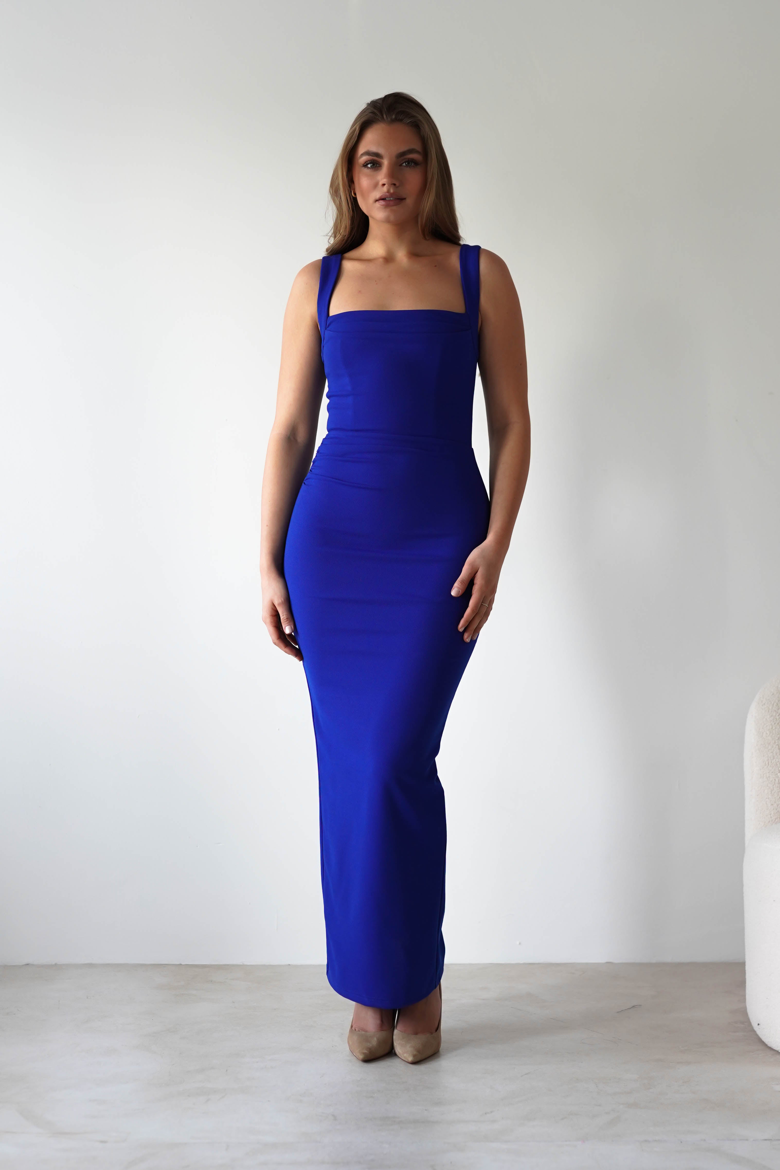 A woman stands against a plain white wall, wearing a royal blue dress with a bodycon fit and beige high heels. Her arms are relaxed at her sides as she faces forward.