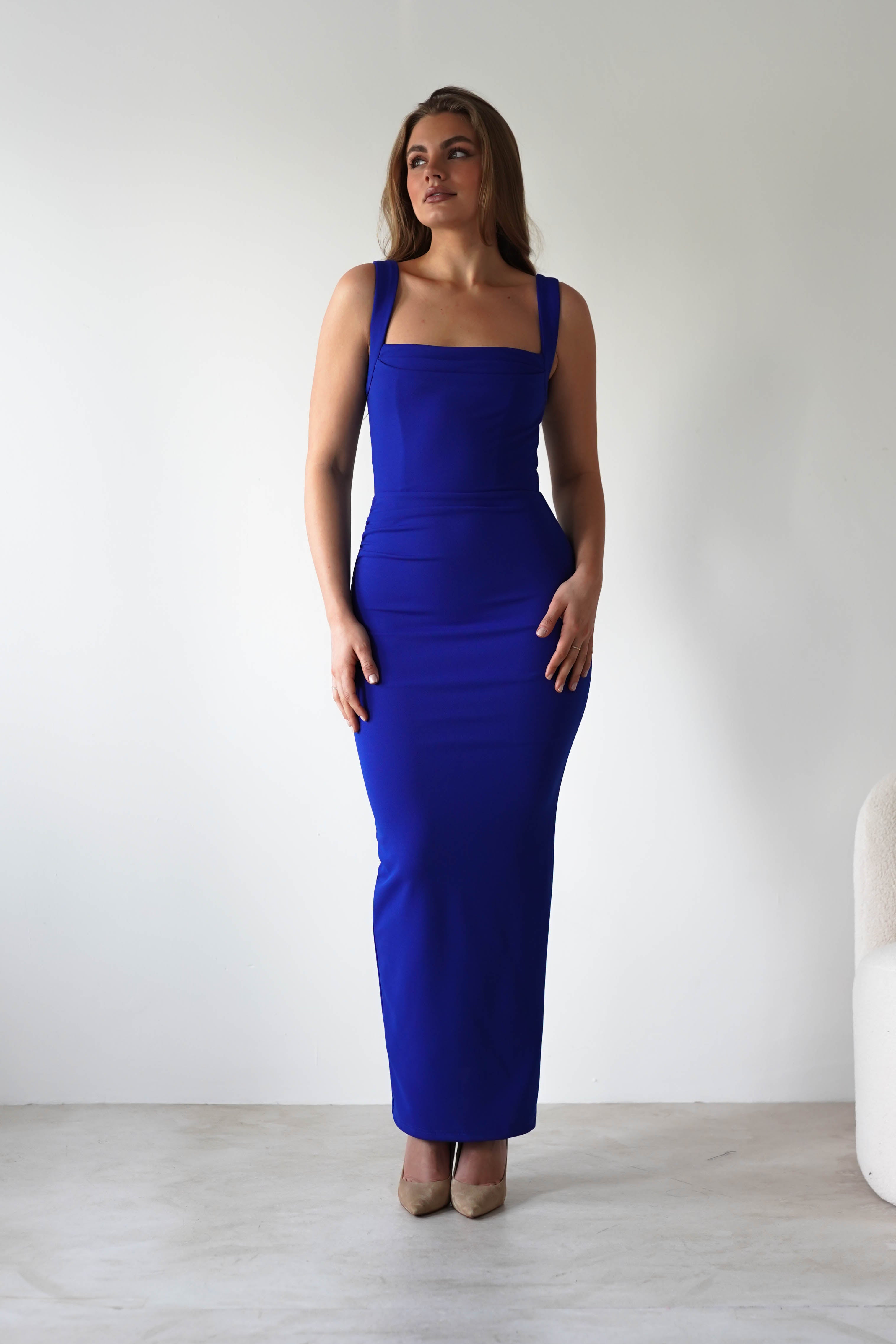 A woman stands against a plain white wall wearing a royal blue, bodycon fit square neck dress. She has light brown hair and beige heels. A white chair is partly visible on the right.