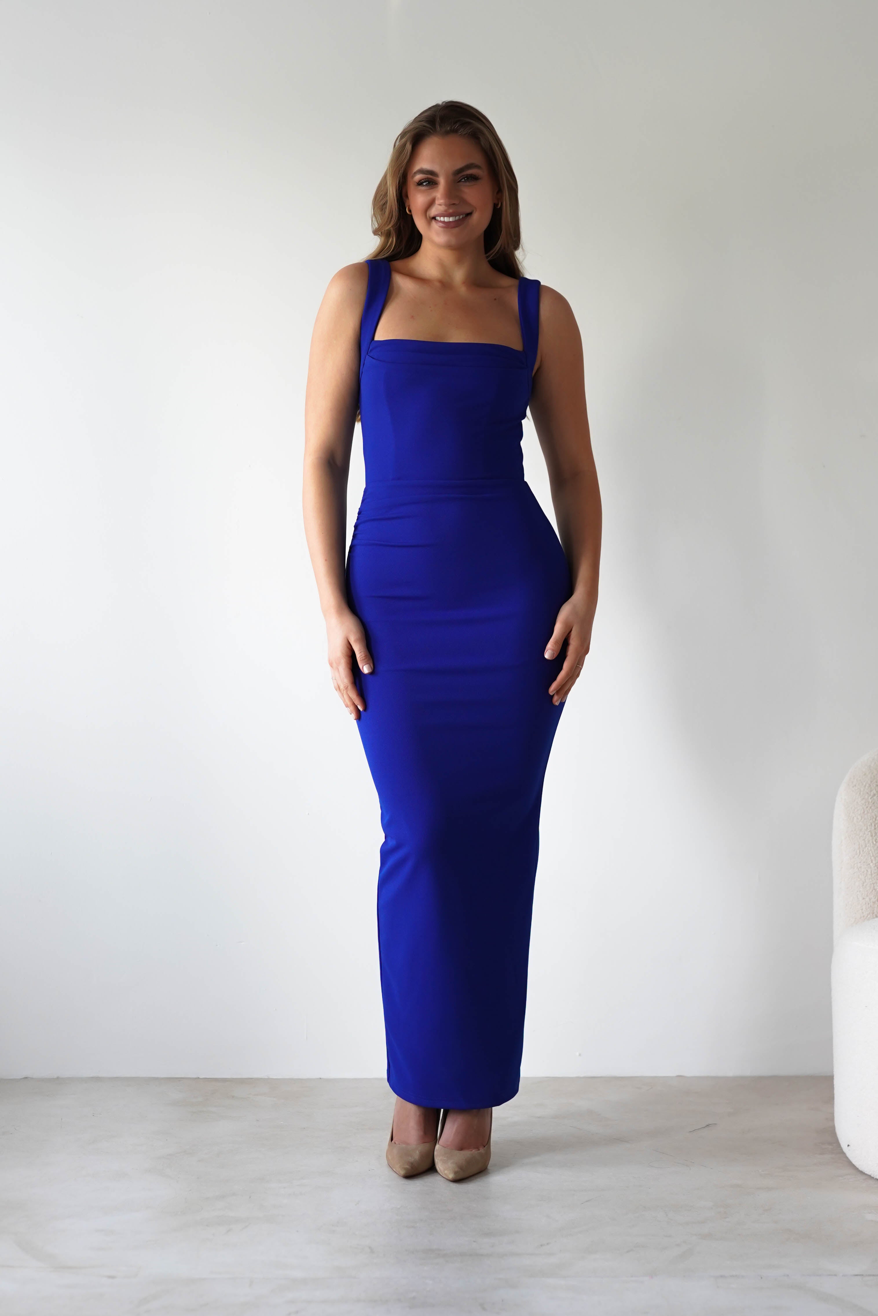 A woman stands smiling in a fitted, royal blue dress with an adjustable tie strap and beige heels against a plain white background next to a white chair.