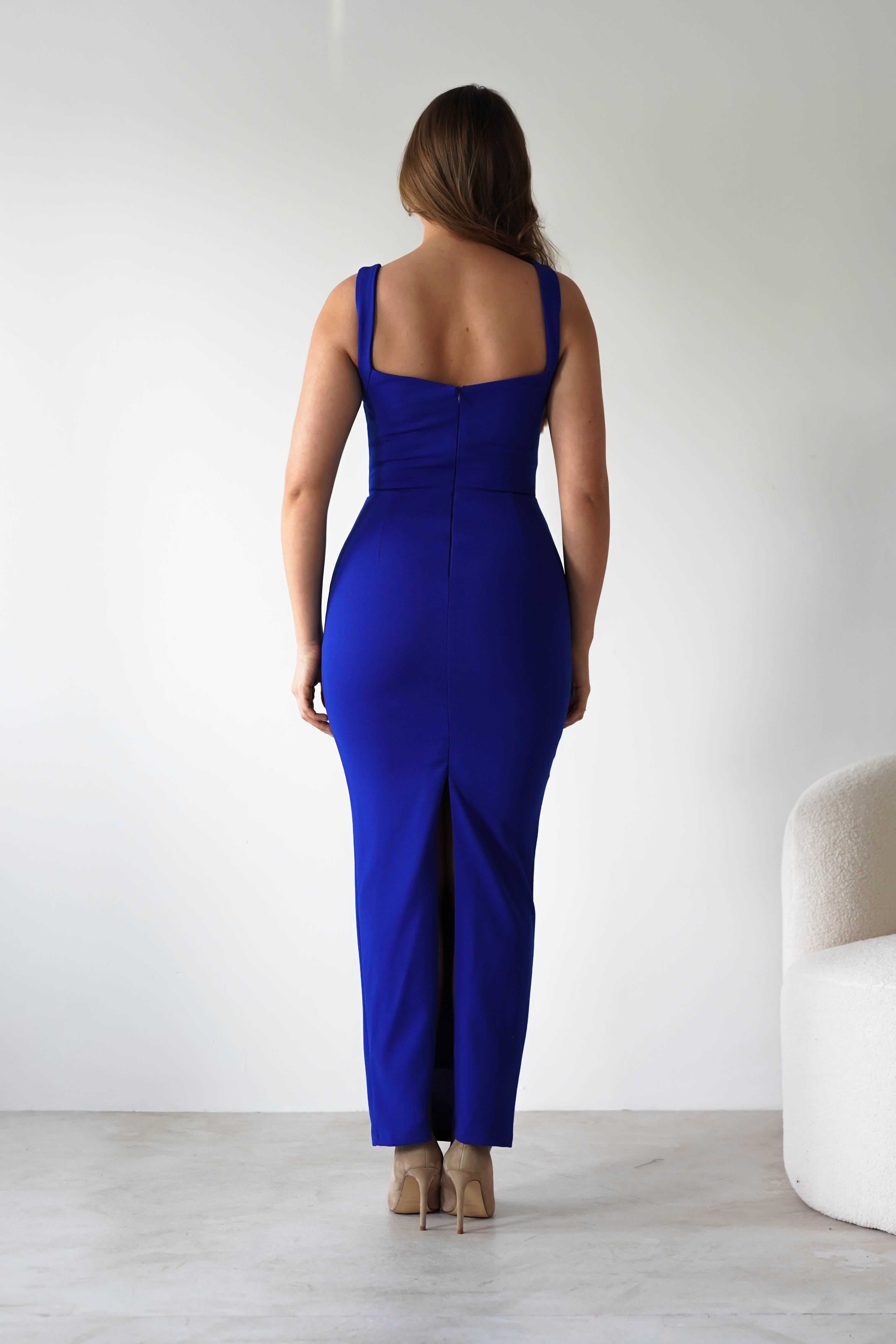 A woman in a fitted, sleeveless royal blue dress with an adjustable tie strap stands facing away in a minimalist room with light walls and a modern off-white chair. She pairs her bodycon fit dress with beige heeled shoes.