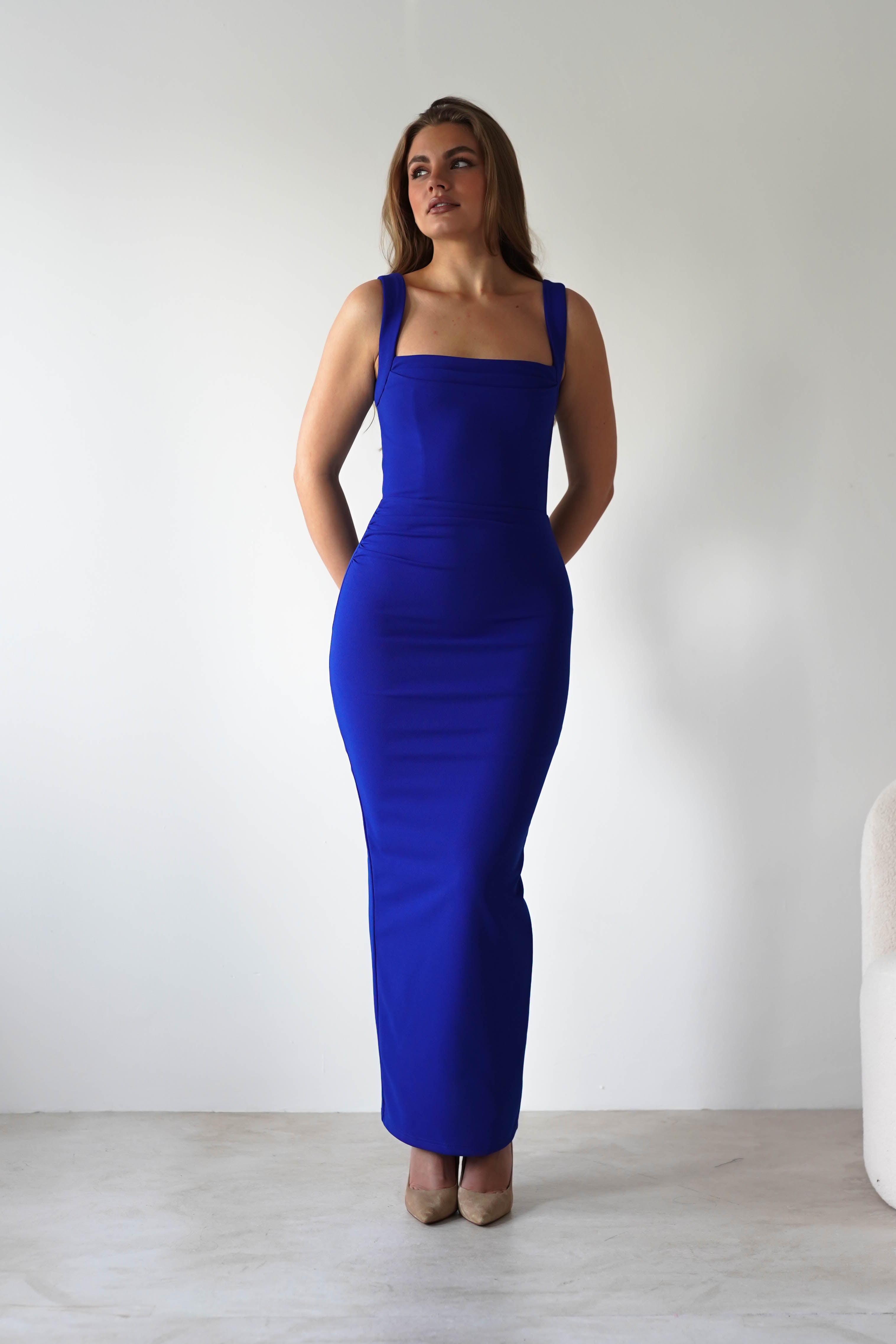 A woman stands indoors against a plain white wall, wearing a royal blue dress with a bodycon fit and beige heels. Her hands are behind her back and she is looking slightly upward.