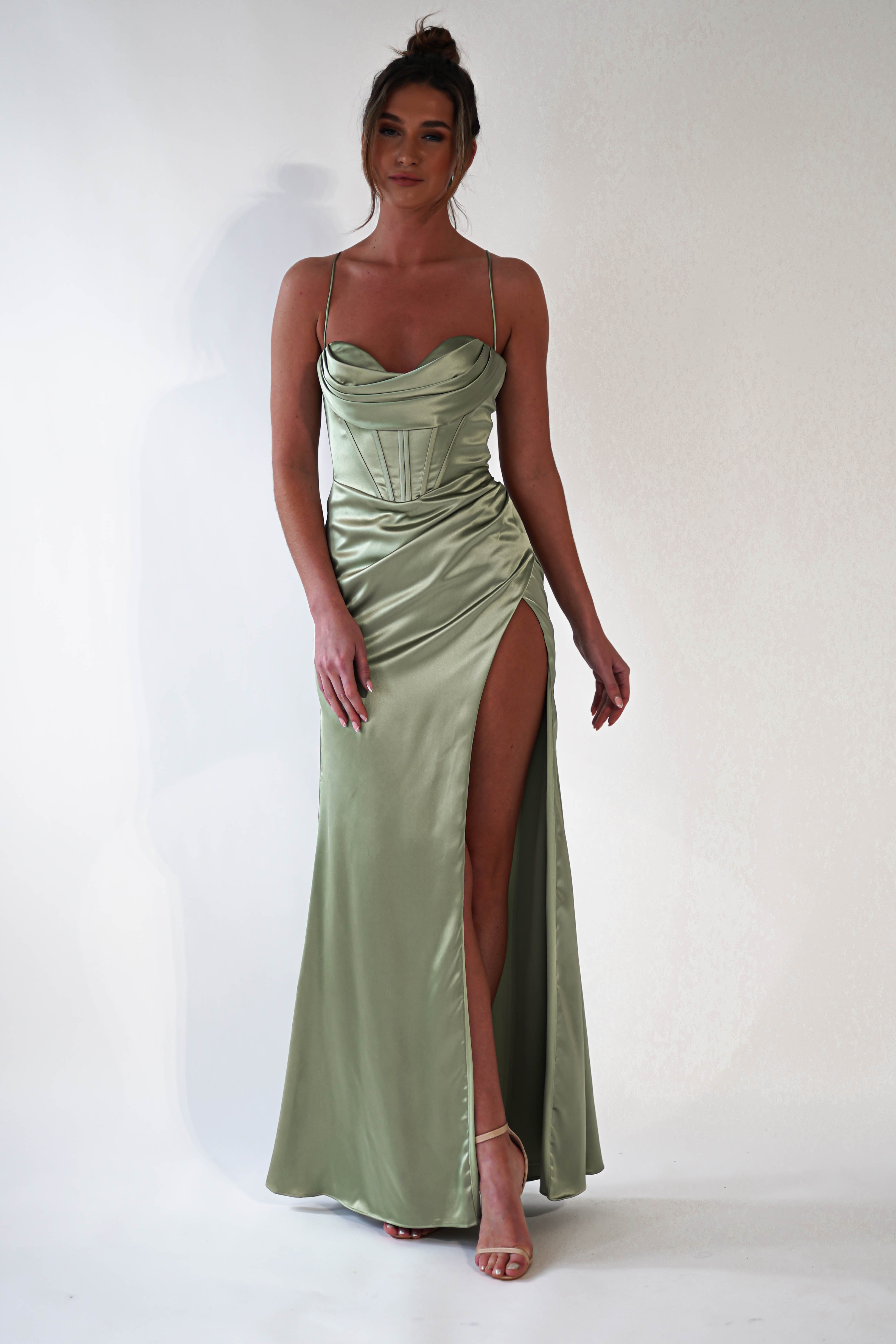 A woman wearing a light green satin corset dress with thin straps and a high thigh slit stands barefoot against a plain white background. With her hair in a loose bun, she looks ready for a special occasion or prom.