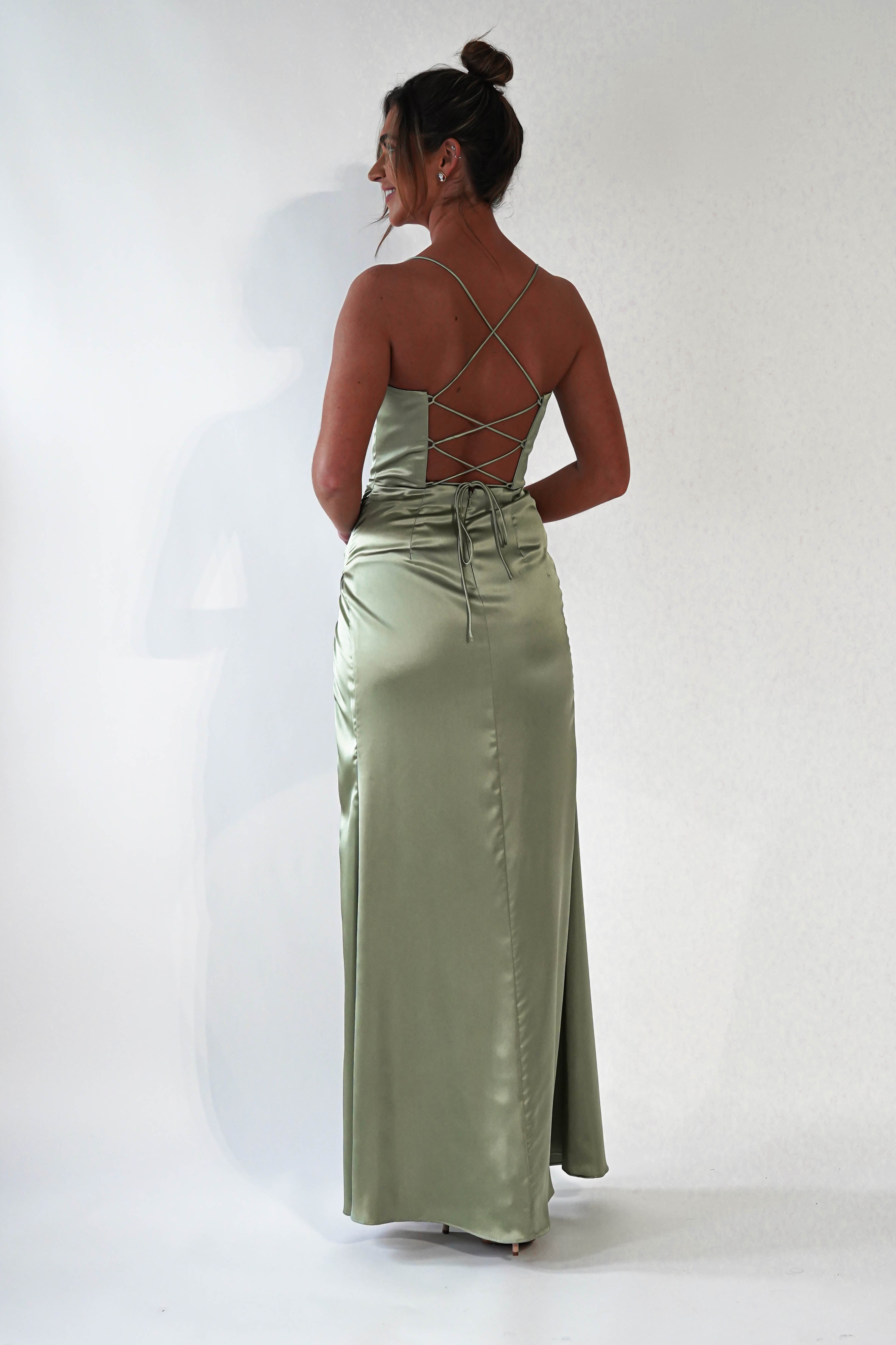 A woman stands facing away, wearing a long, light green satin prom dress with crisscrossed straps on the open back. Her hair is styled in a bun, and she stands against a plain white background.