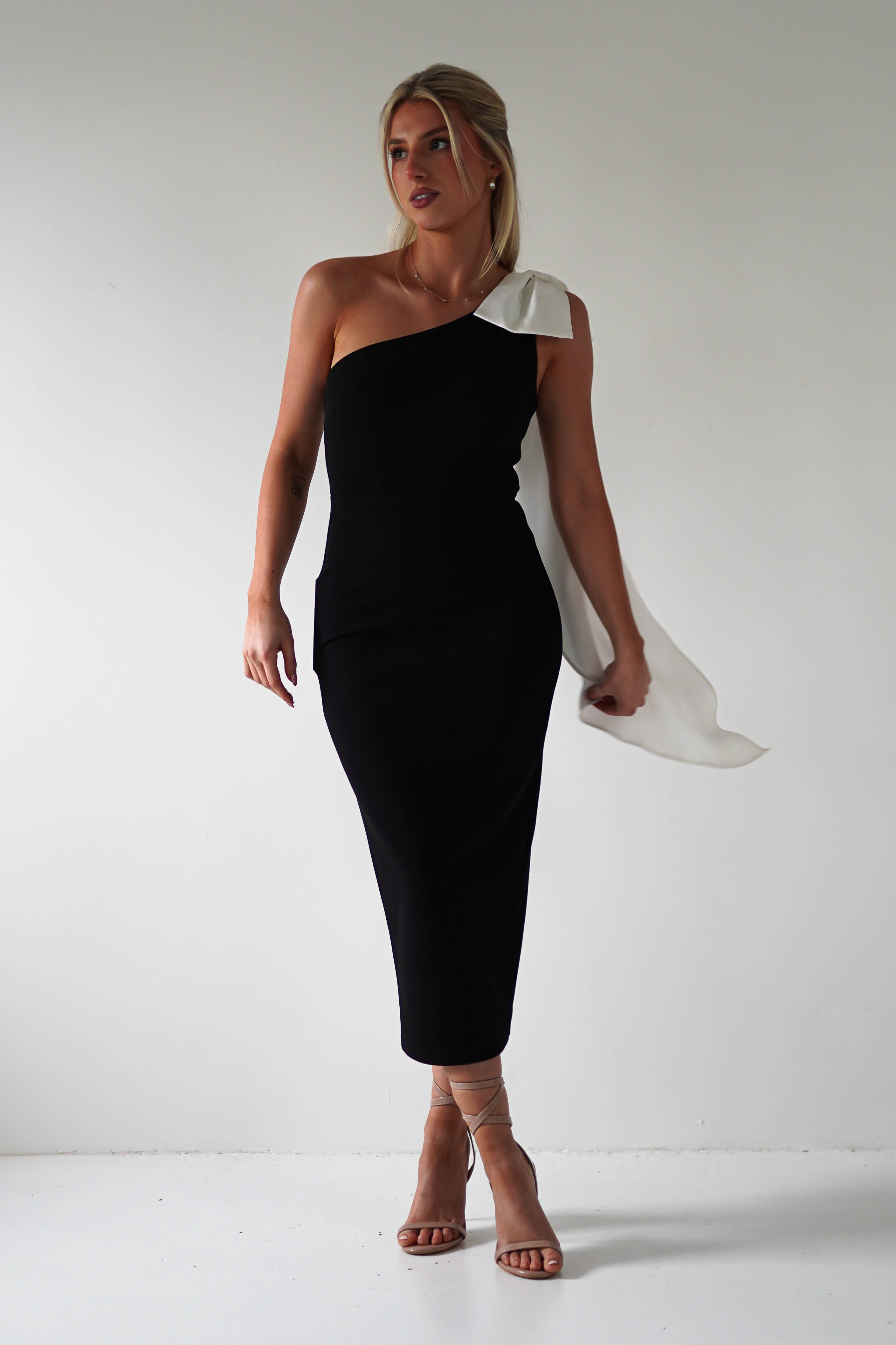 A woman in a black one-shoulder dress, featuring a large white bow, stands against a plain white backdrop. The cute midi dress, made of thick quality fabric, pairs elegantly with strappy beige heels and her styled-up hair—perfect for graduations.