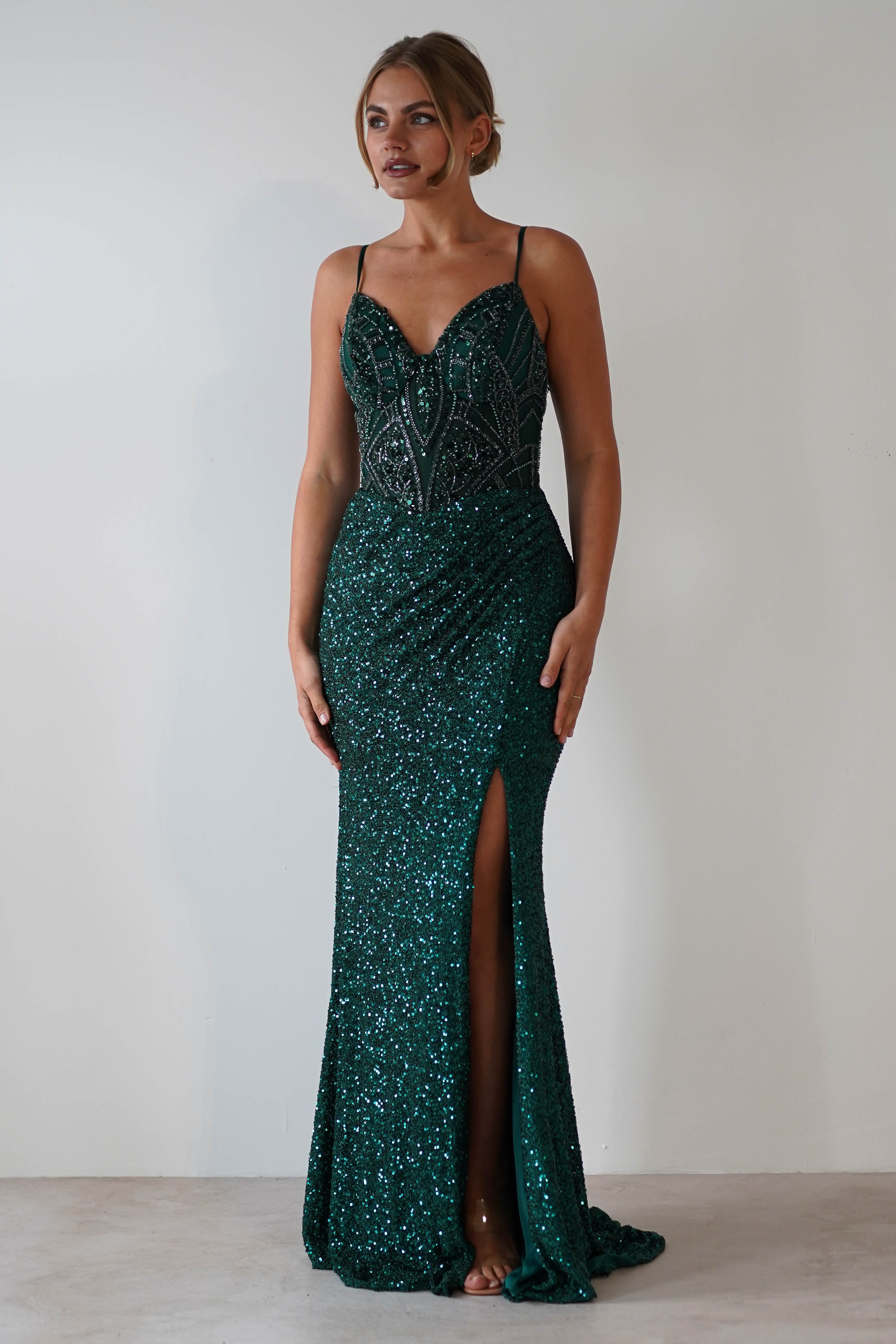 A woman wearing a shimmering emerald green mermaid gown with thin straps, intricate beading on the bodice, and a thigh-high slit stands against a plain light-colored background.