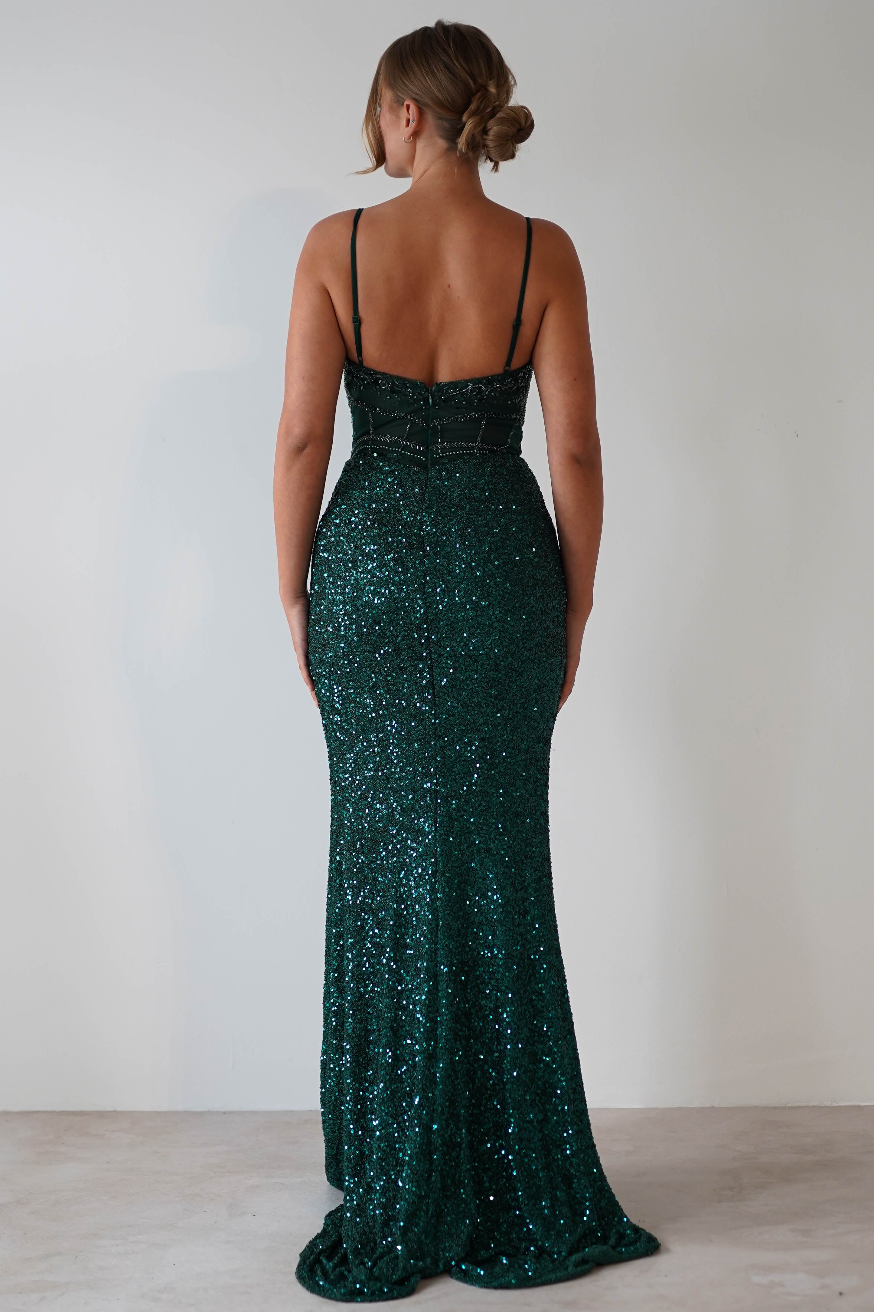 A woman with blonde hair in a low bun stands with her back to the camera, wearing a dark green, floor-length sequin dress with thin straps—a stunning mermaid gown perfect for prom—against a plain light-colored background.