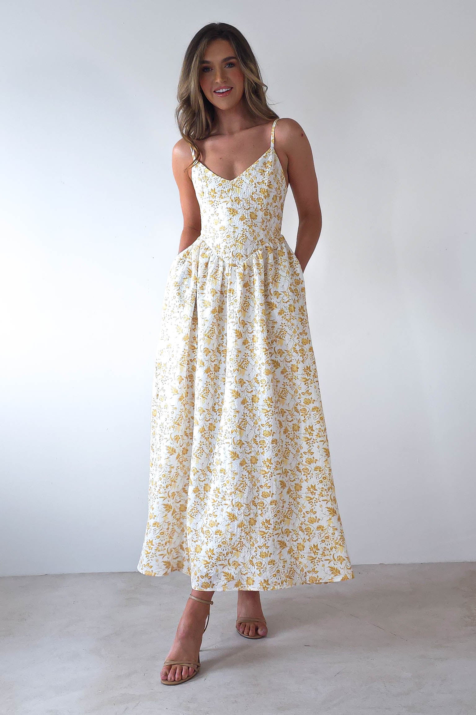 A woman in a sleeveless summer print maxi dress stands against a plain background. The dress, ideal for wedding guests, is ankle-length with a flowing skirt and adjustable ties. She has long hair, wears strappy sandals, and smiles with hands in pockets.