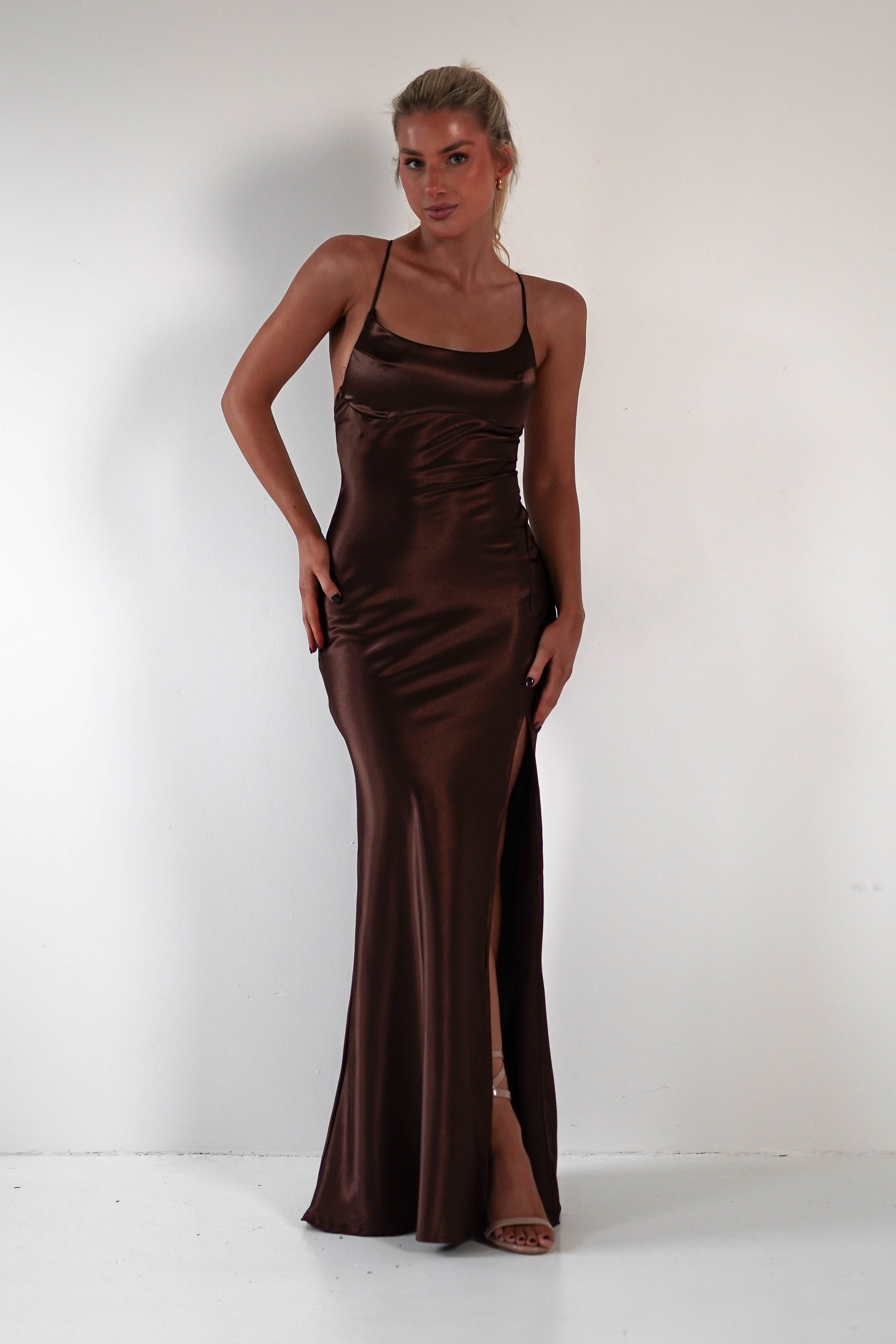 A woman in a sleek, chocolate brown satin gown stands against a white background. The prom dress features thin straps and a high slit, accentuating the figure-hugging silhouette. She poses confidently, looking towards the camera, with her blonde hair pulled back.