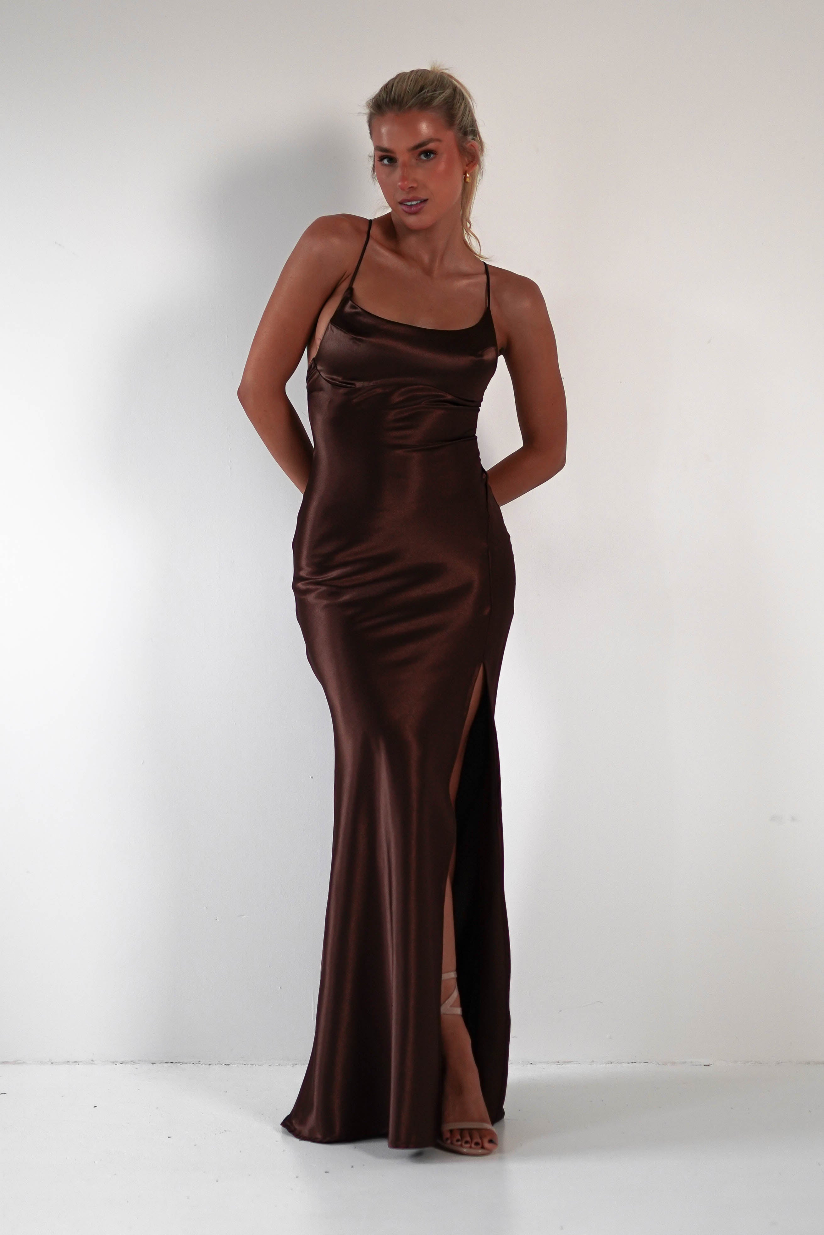 A woman in a sleek, brown satin gown with spaghetti straps stands against a plain white background. The figure-hugging silhouette of the dress features a side slit, and she poses with her hands behind her back, looking confidently at the camera.