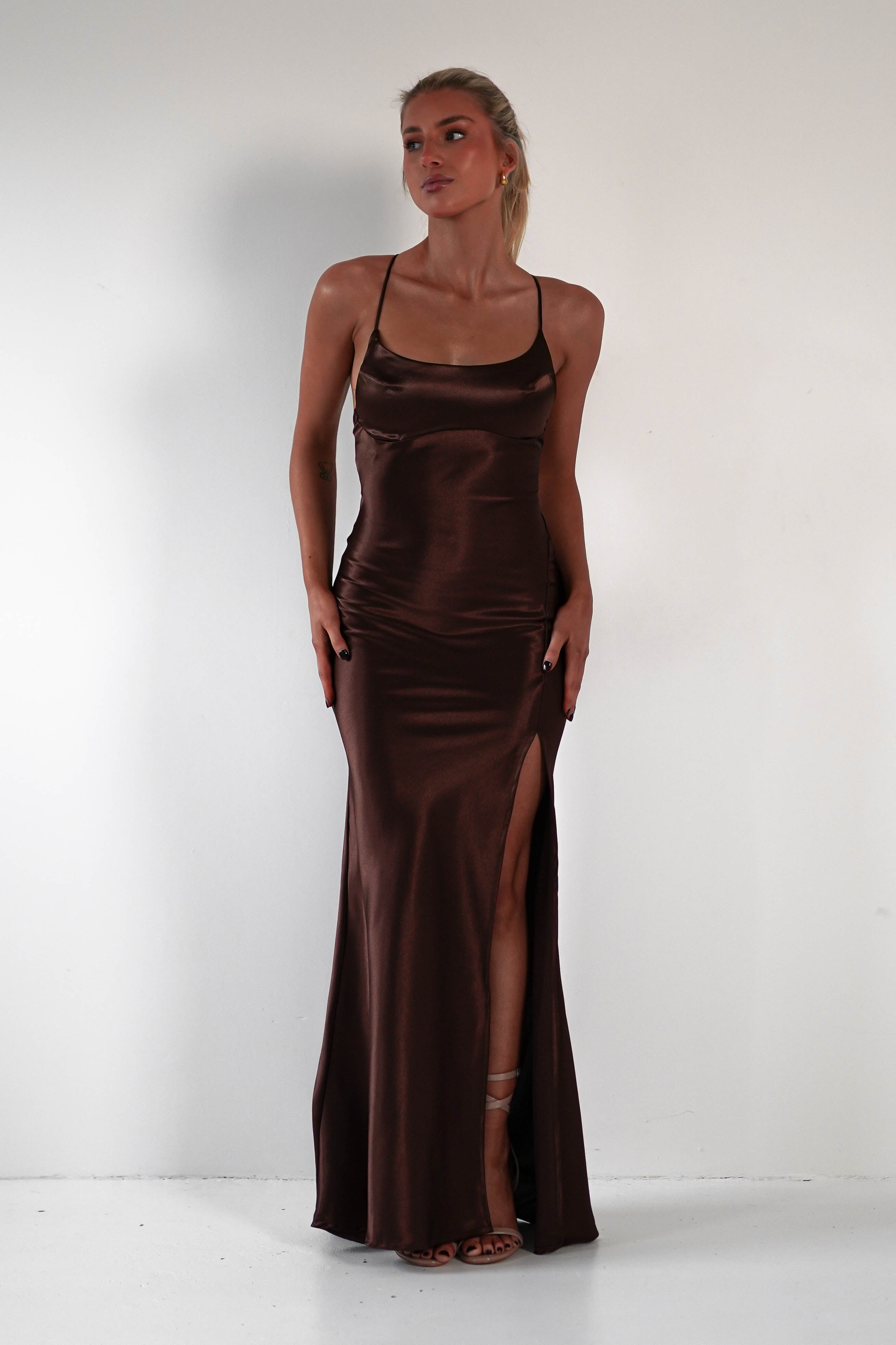 A person is wearing a long, sleek brown satin gown with thin straps and a thigh-high slit. They have long hair tied back and are posing in front of a plain white wall. The figure-hugging silhouette is completed with strappy sandals.