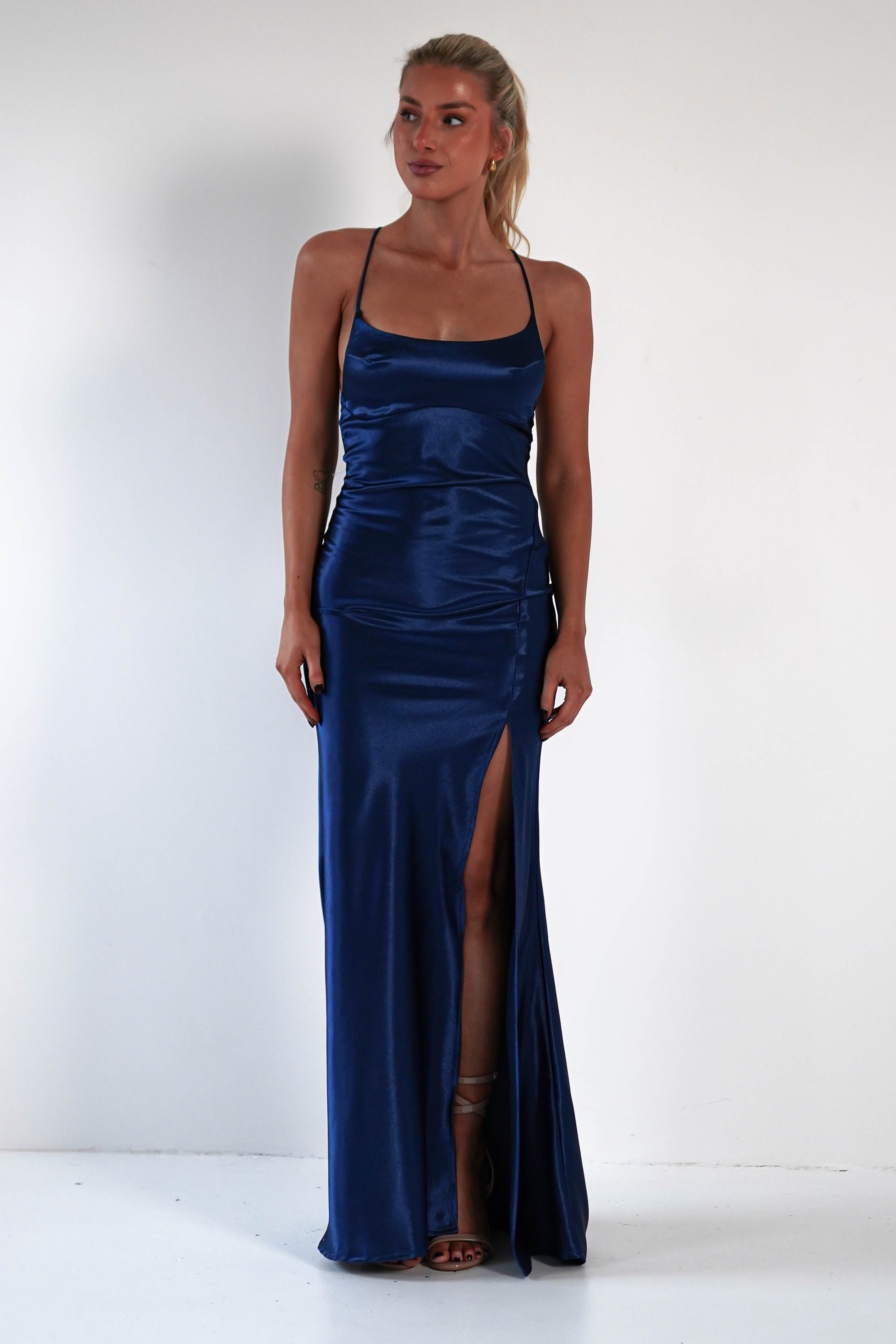 A woman wearing a floor-length, royal blue satin gown with thin straps, a figure-hugging silhouette, and a high slit on one side stands against a plain white background.