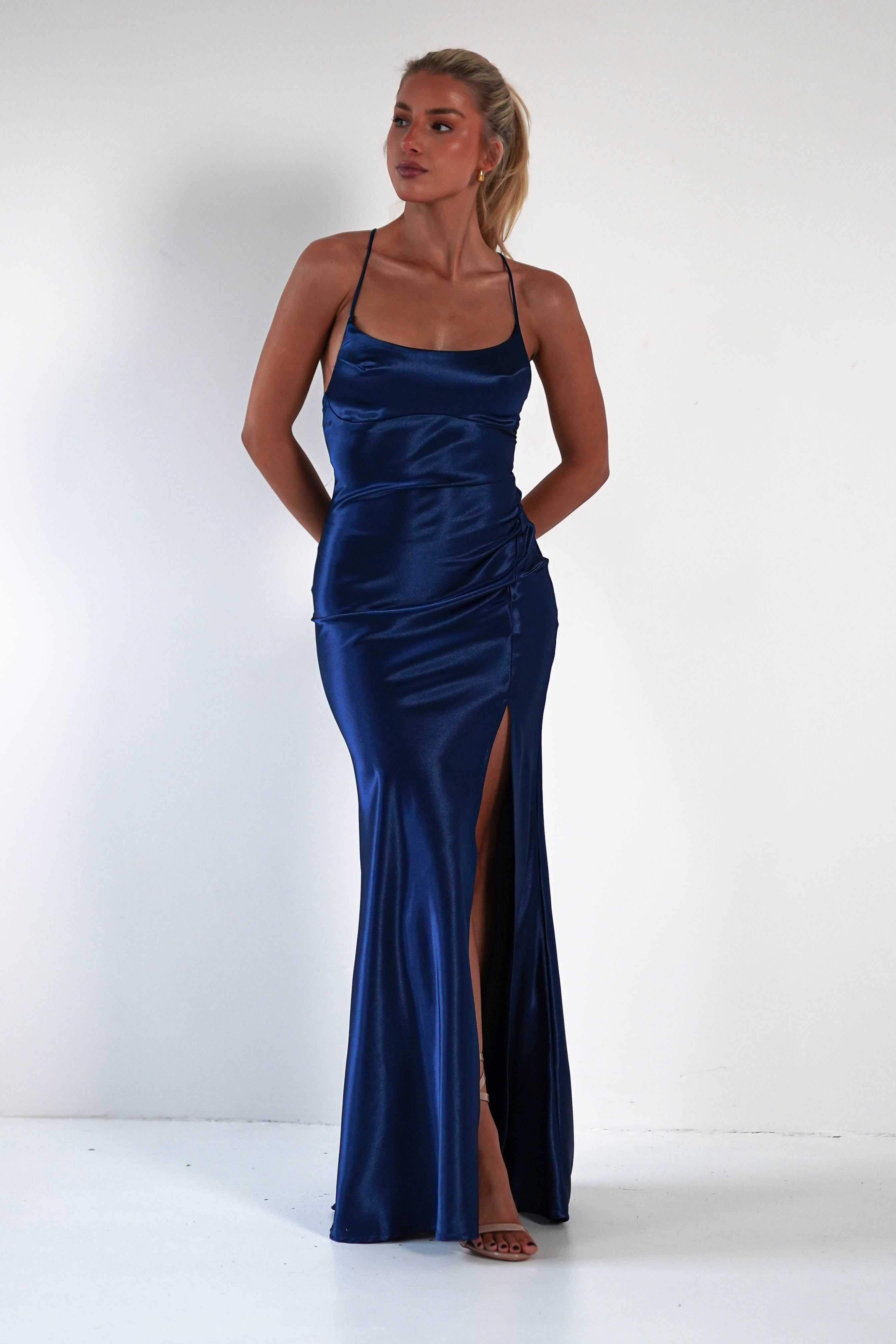 A woman stands against a plain white background in a royal blue satin gown with thin straps, a high front slit, and a figure-hugging silhouette. She has her hands by her sides and gazes slightly to the left.