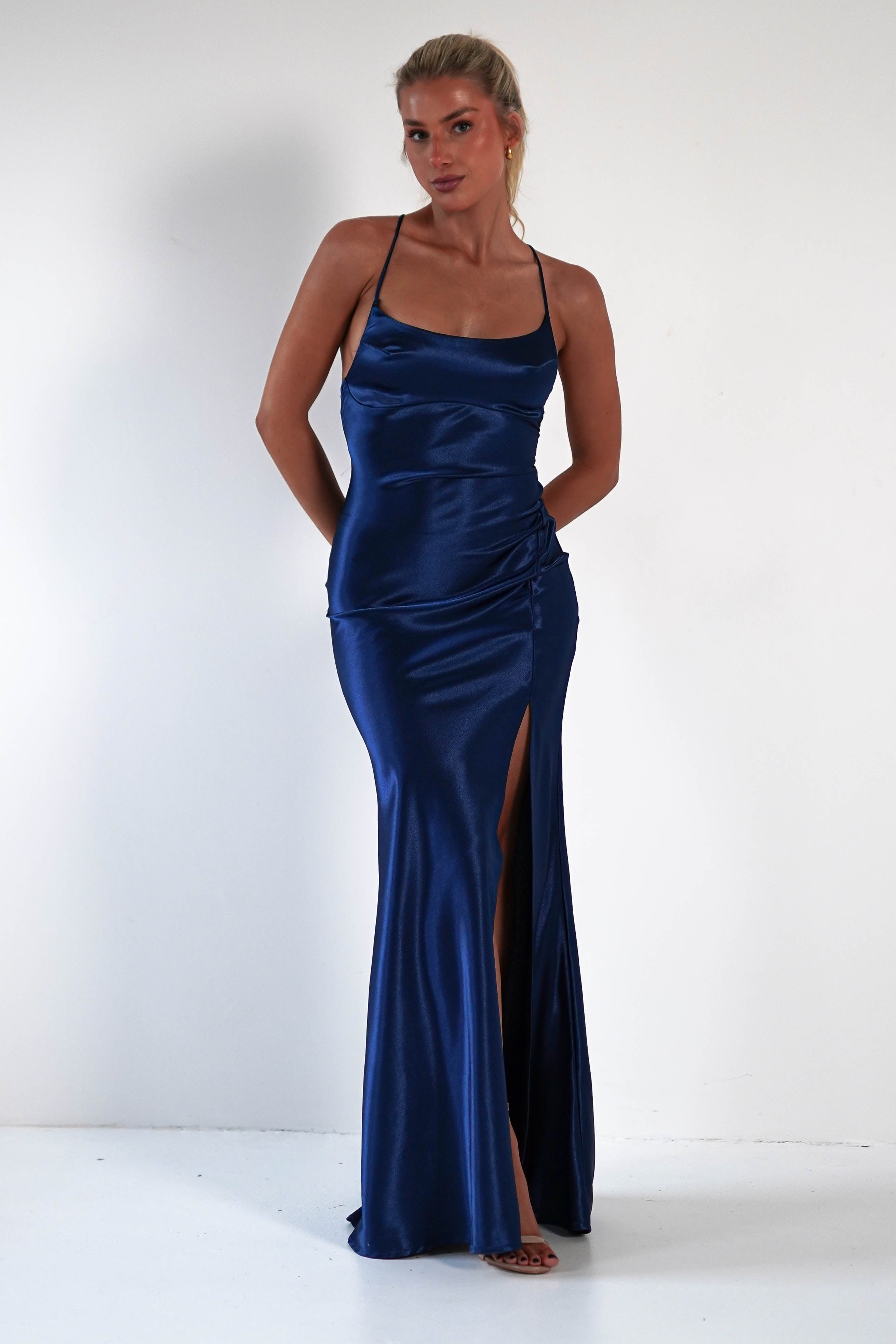 A woman wearing a royal blue satin gown with a figure-hugging silhouette, sleeveless design, thin straps, and a high thigh slit stands against a plain white background.