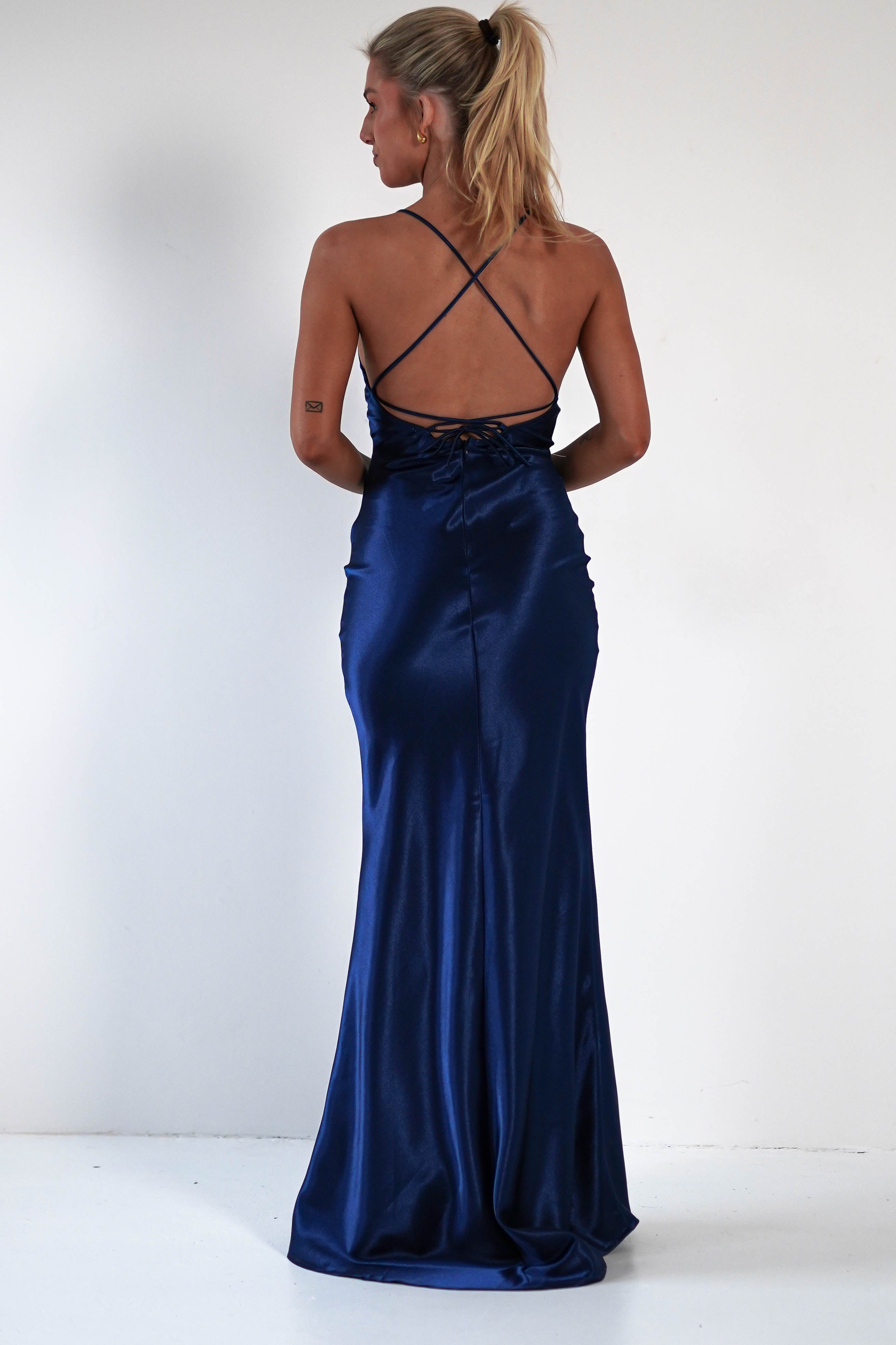 A woman with blonde hair in a ponytail wears a royal blue satin gown with a low, open back and crisscross straps, showcasing a figure-hugging silhouette as she stands against a plain white wall.