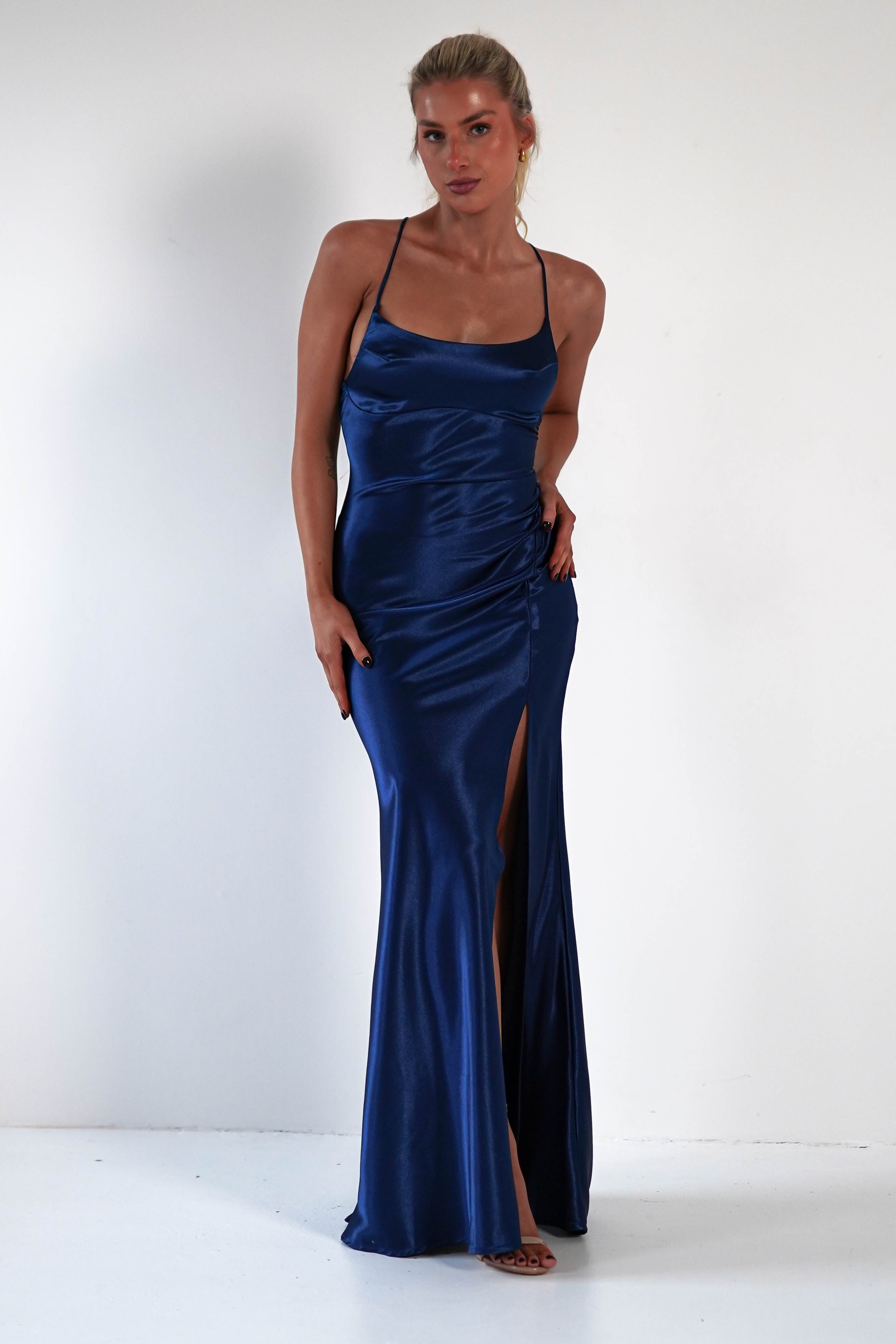 A woman poses in a sleeveless, floor-length royal blue satin gown with thin straps and a thigh-high slit, accentuating the figure-hugging silhouette as she stands against a plain white background.