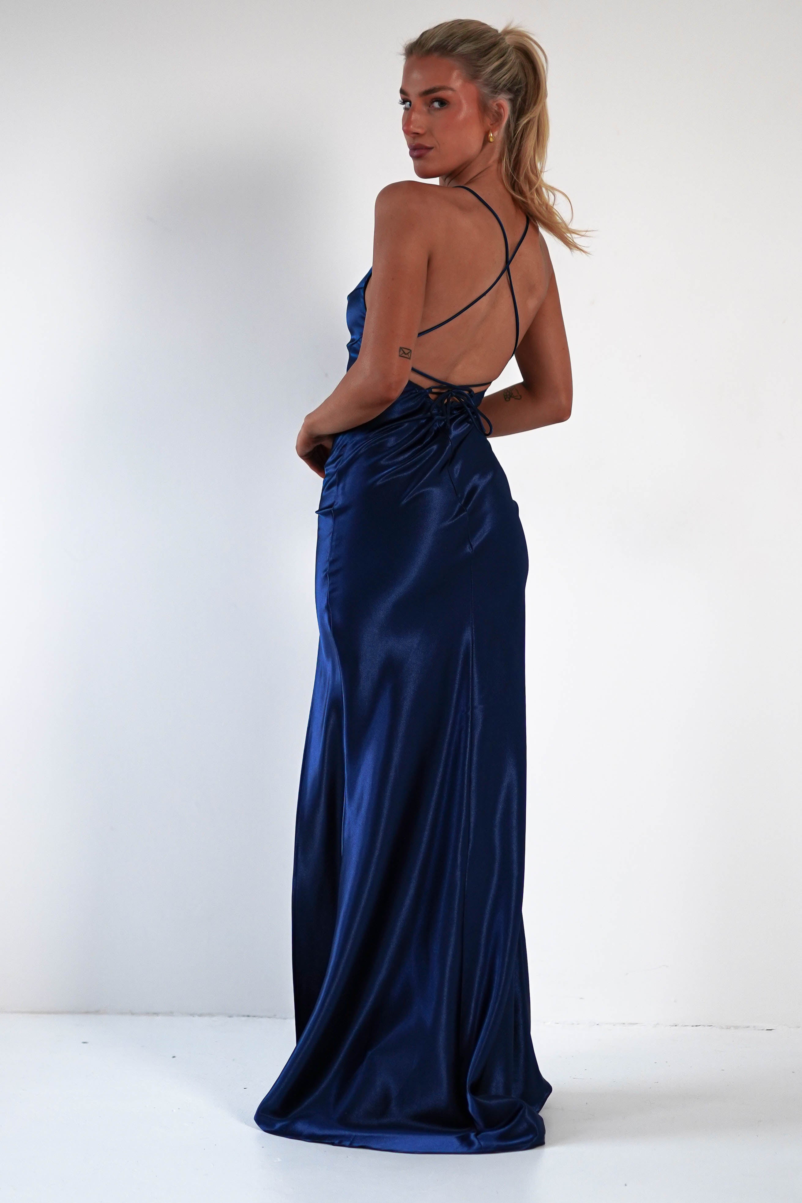A woman stands in a figure-hugging, backless royal blue satin gown with thin crisscross straps, looking over her shoulder. Her blonde hair is in a ponytail as she poses against a plain white background.