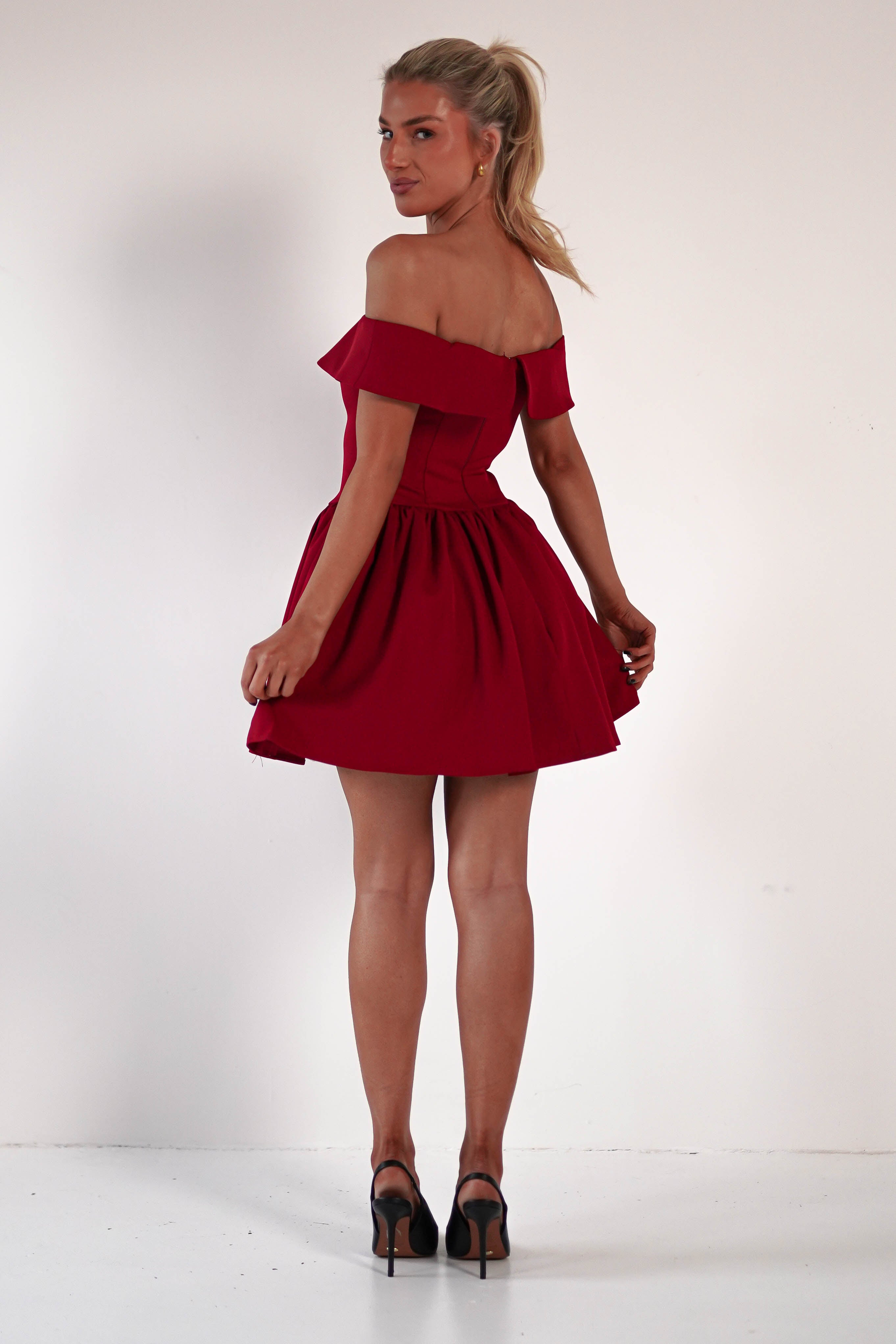 A person in a red, off-the-shoulder skater mini dress and black high heels stands against a plain white background, looking back over their shoulder. Their hair is styled in a ponytail.