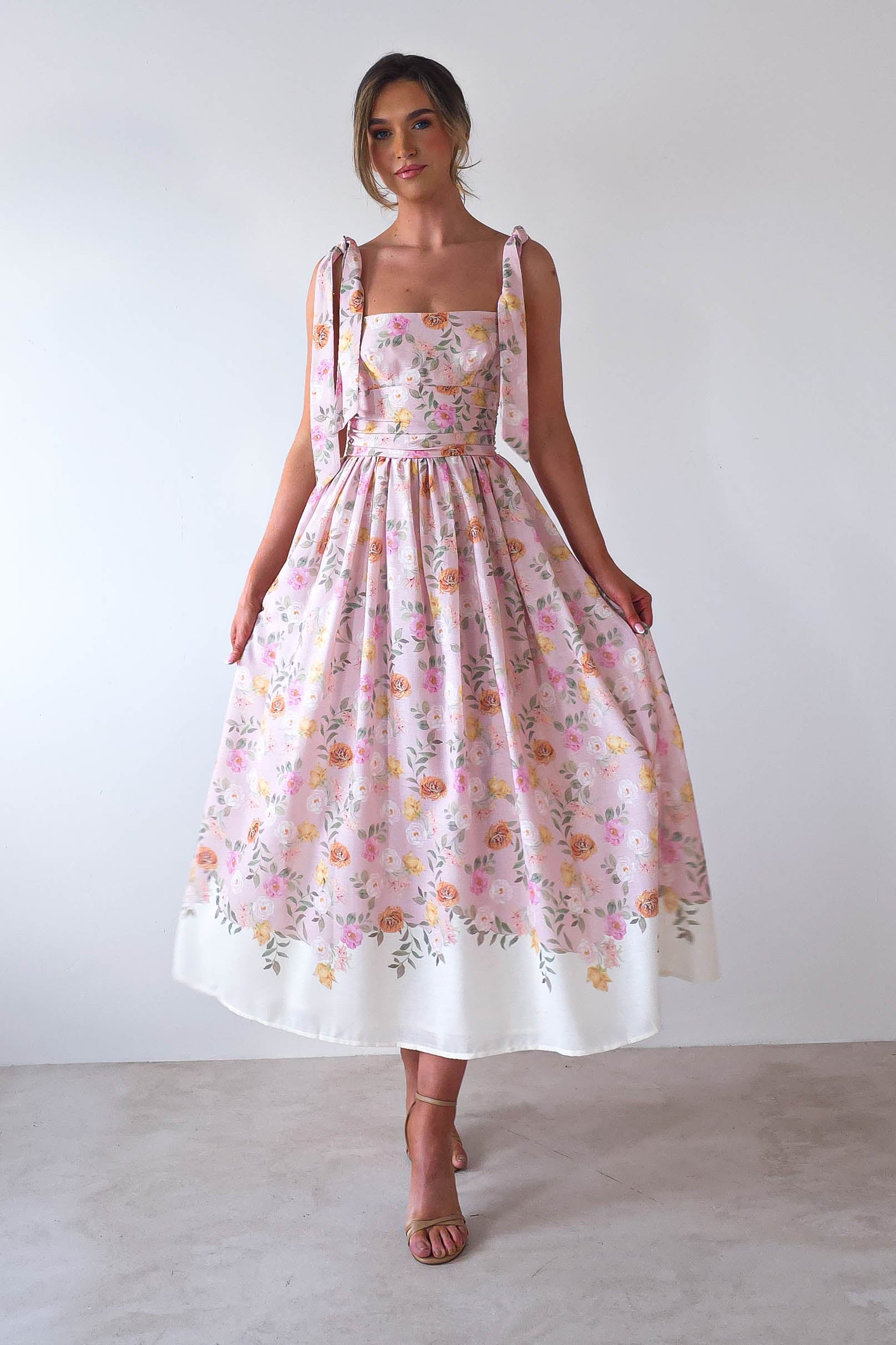 A woman stands gracefully in a sleeveless, floral print pink dress with tied shoulder straps—perfect for summer events. The full skirt elegantly drapes over her brown heels against a plain white background, capturing the essence of a charming summer print maxi dress.