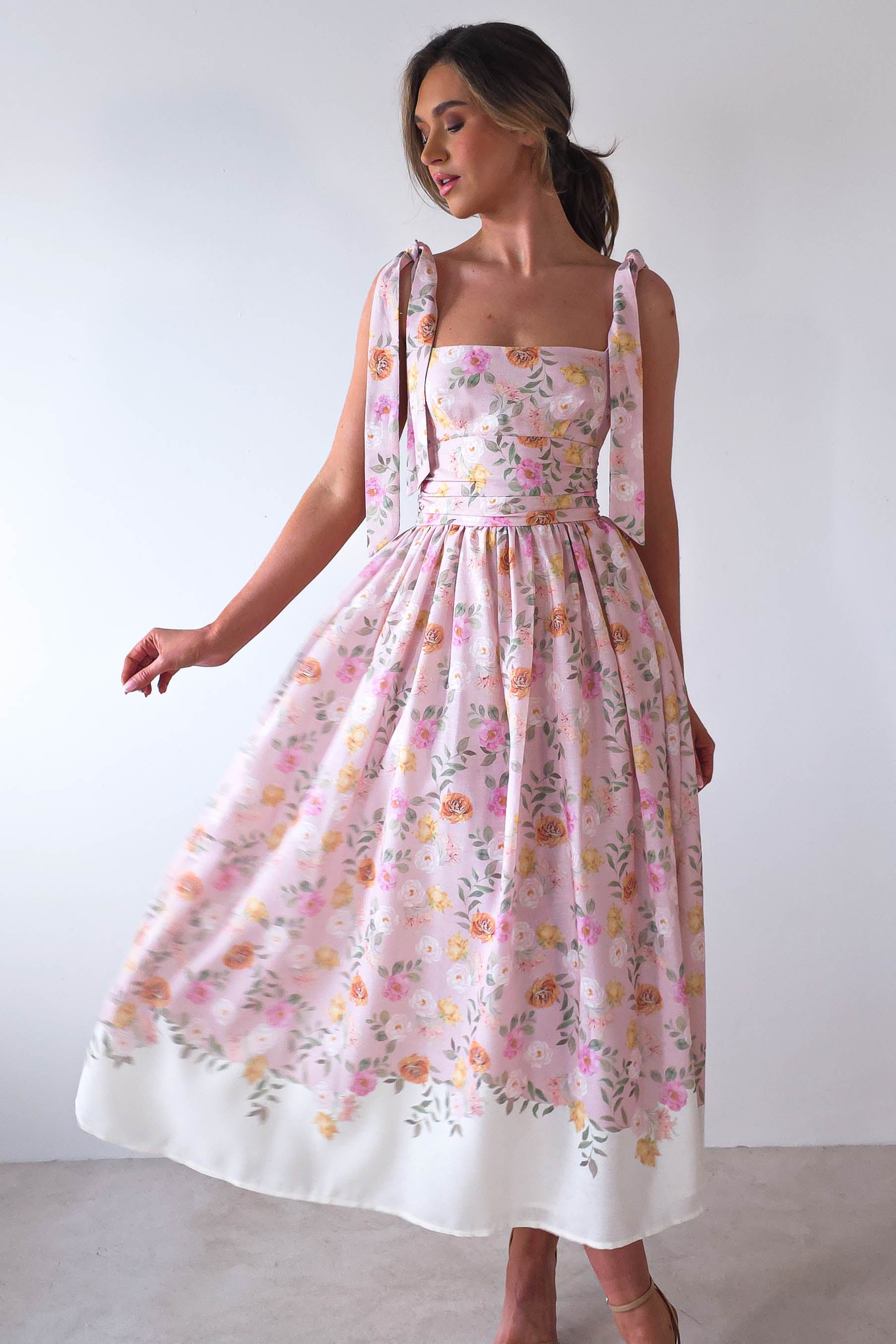 A person poses in a flowy, sleeveless pastel floral dress with a fitted bodice and full skirt, perfect for summer events. The dress has wide shoulder straps tied in bows, and the person stands against a plain light-colored background.