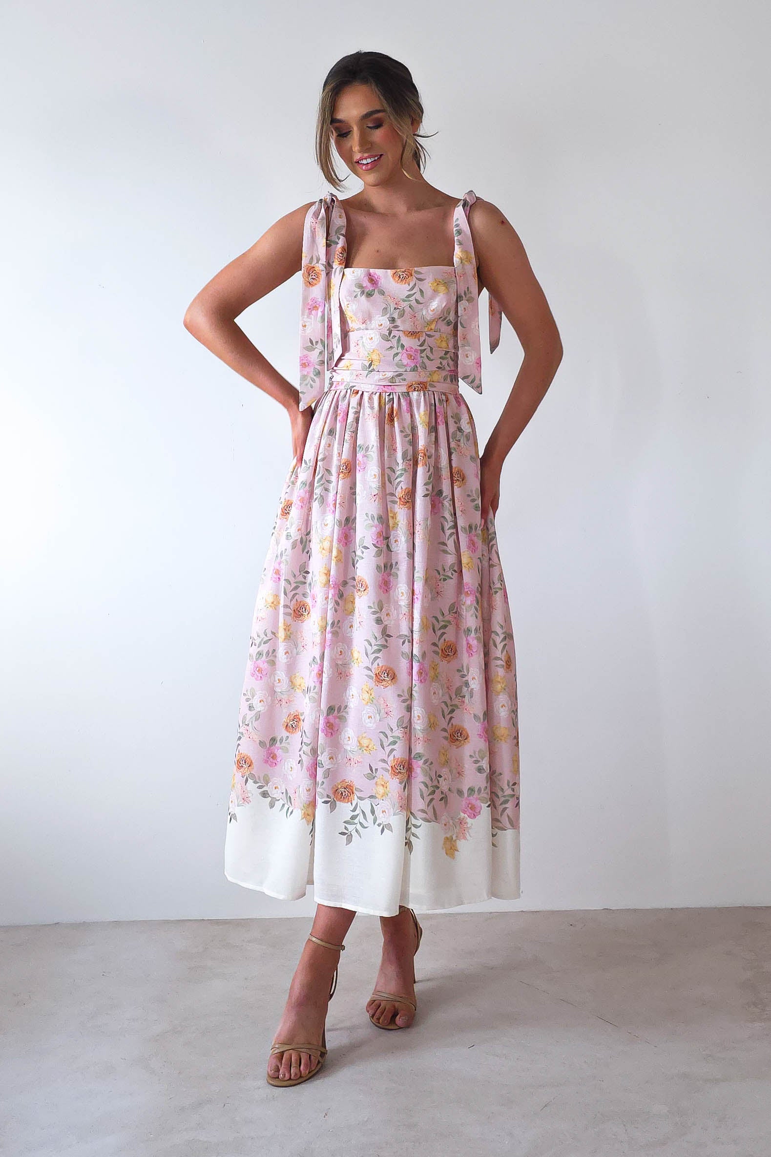 A person wearing a sleeveless, summer print maxi dress with shoulder tie straps stands against a plain white background. Perfect for wedding guests or summer events, they smile confidently with hands on their waist, complemented by light-colored heels.