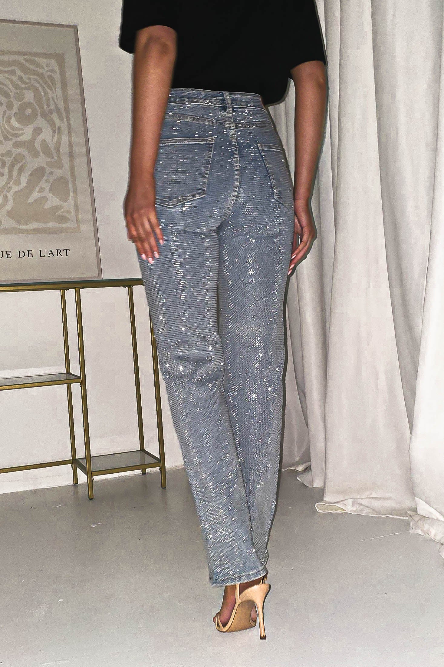 A person in a black top and Diamante Sparkle Jeans stands indoors facing away. They are wearing beige high-heeled sandals. A framed artwork and a metal shelf are visible against a light-colored wall and drapes, accentuating the rhinestone embellishment of their high-quality denim attire.