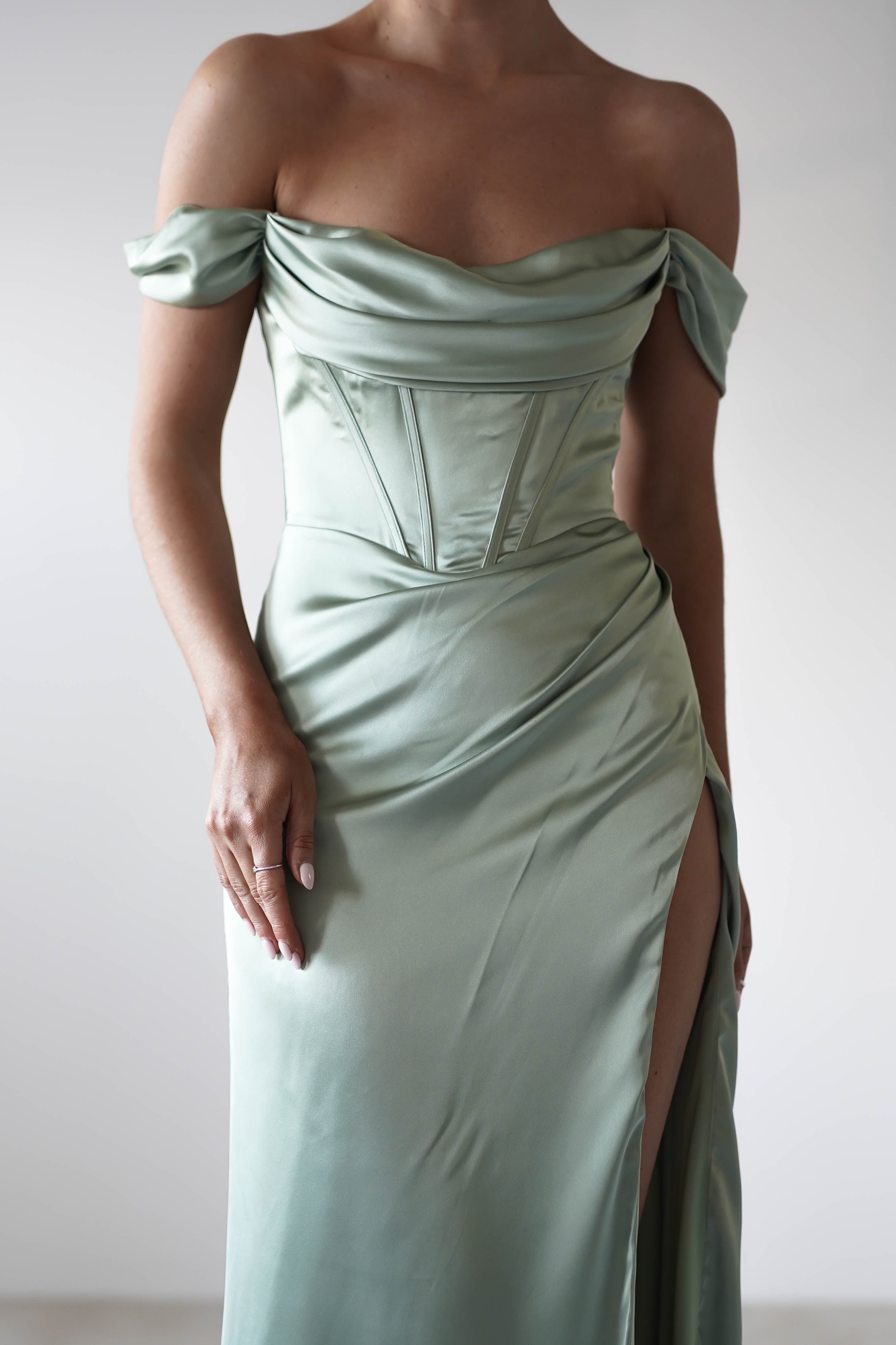 A person wears an elegant, off-the-shoulder, light green satin dress with a corset-style bodice and high slit. Crafted from thick satin fabric, the image is cropped at the neck and mid-thigh.