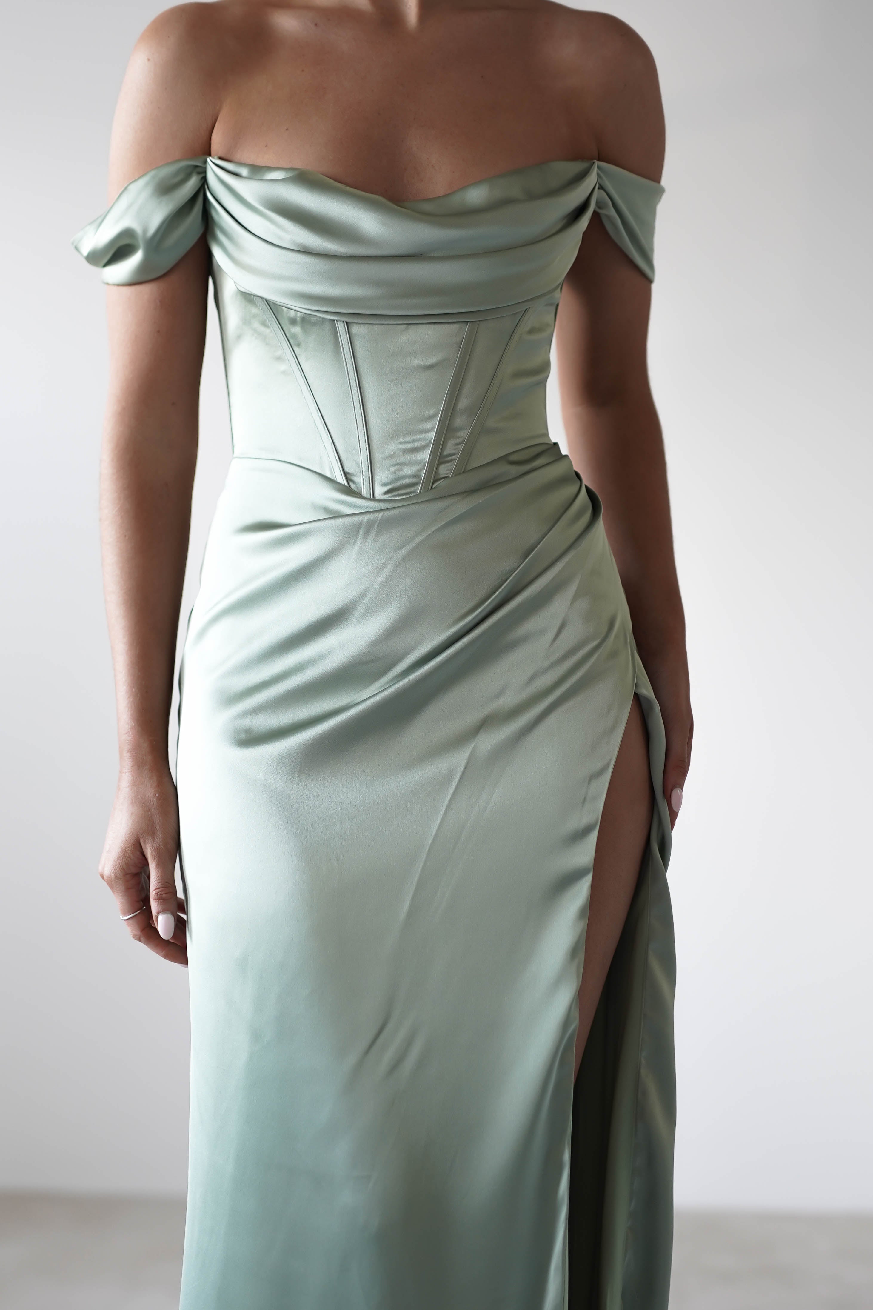 A person wearing an elegant, light green off-the-shoulder satin dress with a fitted corset style bodice and a high slit on one side, crafted from thick satin fabric, stands against a plain light background.