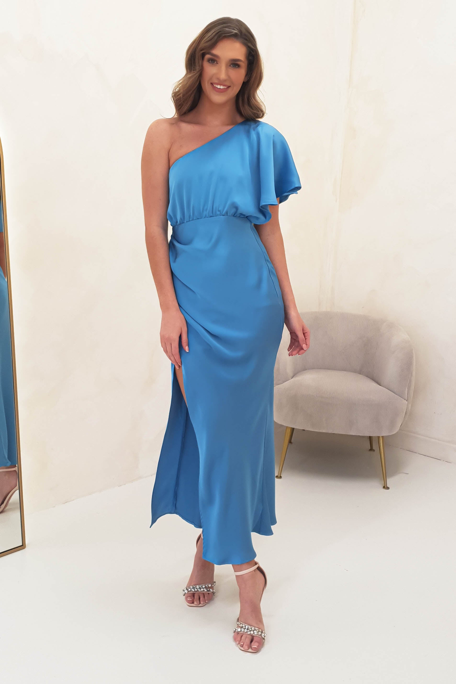 A woman in a one-shoulder satin maxi dress with a high slit stands smiling indoors next to a light beige armchair. She has wavy hair, strappy heels, and could easily inspire bridesmaids looks with her elegant style. The background is simple and light-colored.