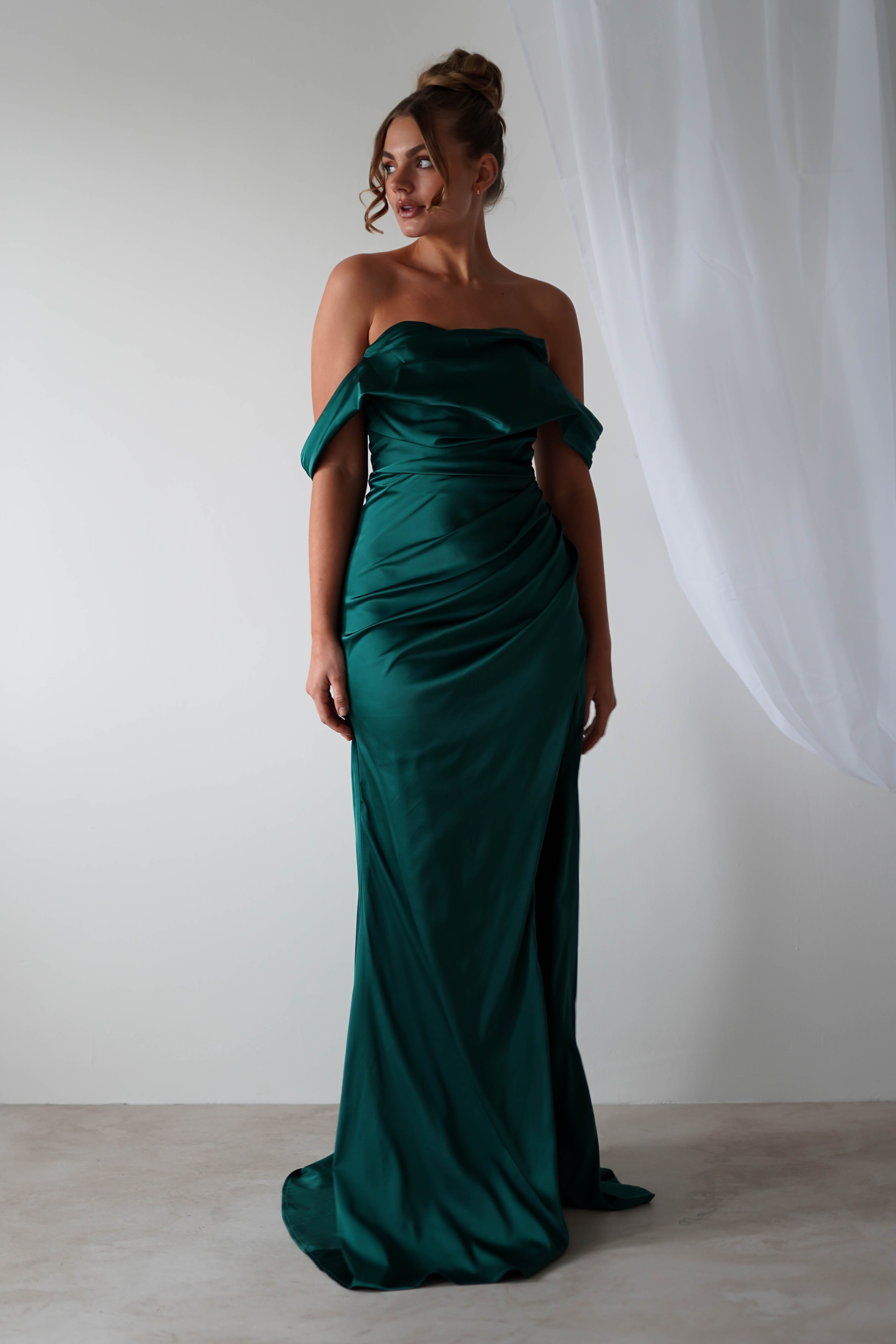 A woman stands in a green, off-the-shoulder gown made from satin material with gathered fabric details. Her hair is styled in an updo as she poses against a plain white background with sheer white curtains—perfect for bridesmaids.