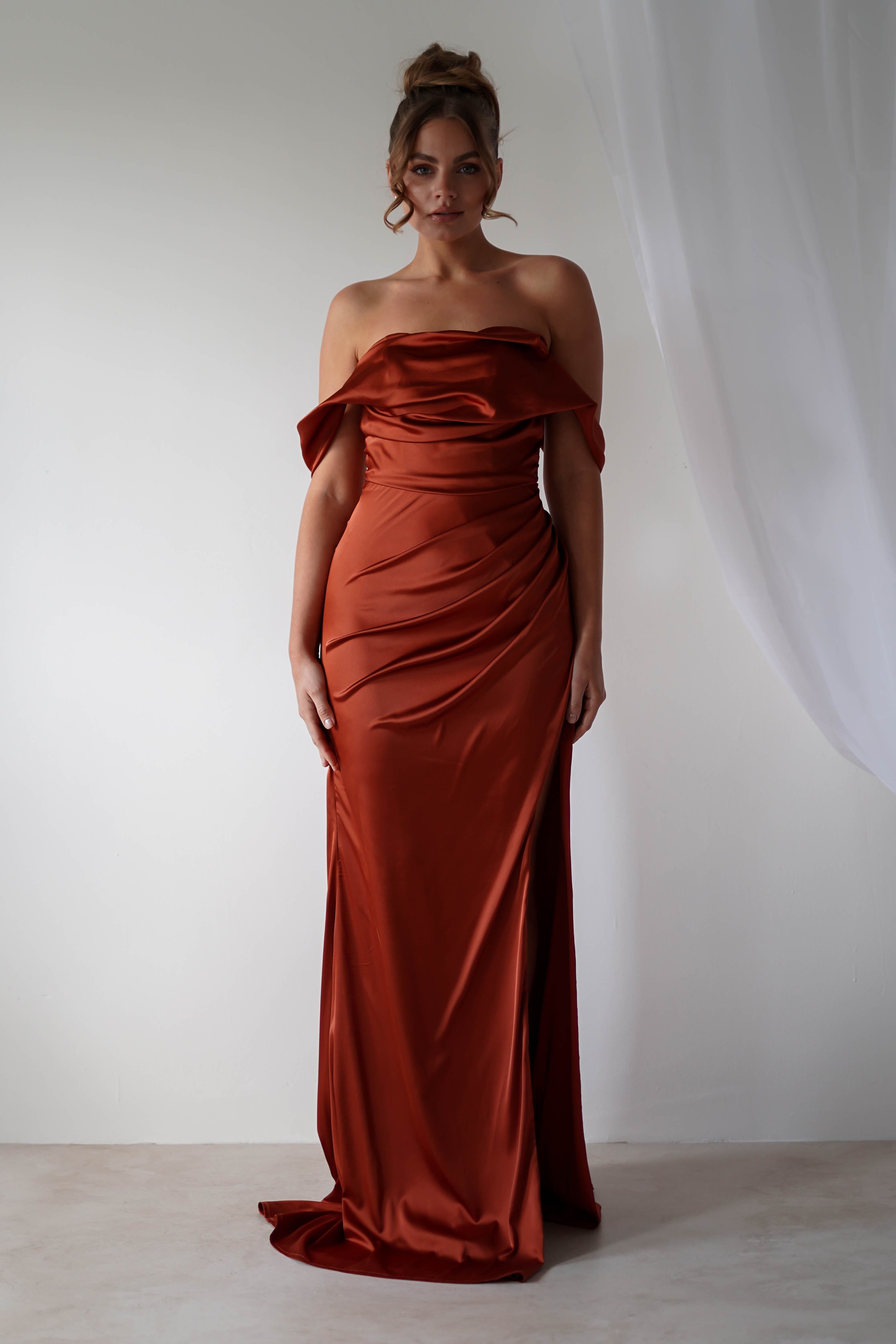 A woman stands in front of a white background wearing an off-the-shoulder gown crafted from rust-colored satin material, featuring a fitted, draped silhouette and a small train—perfect for formal events. Light sheer curtains hang to the right.