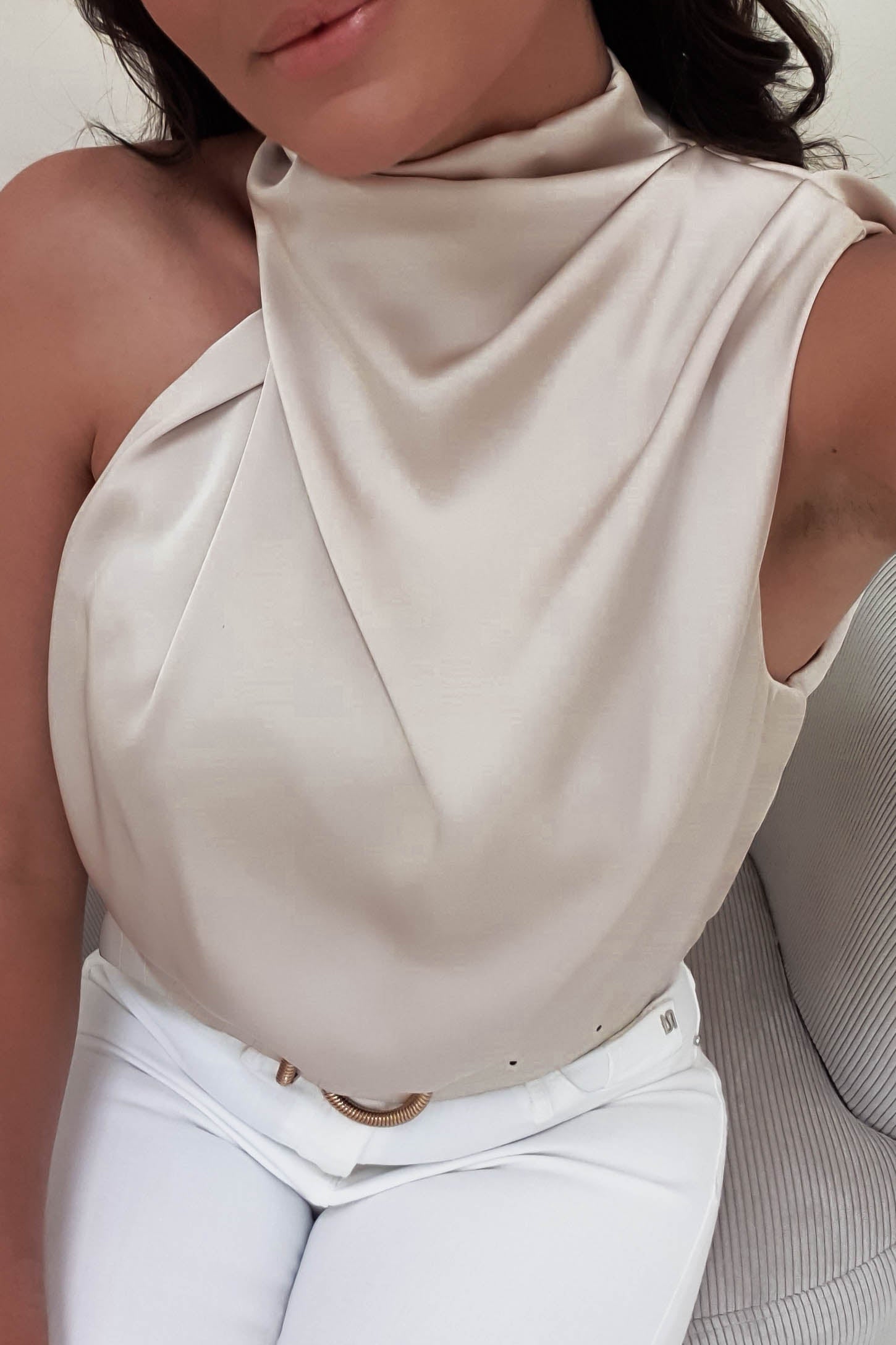 A woman wearing a stylish, cream-colored, draped halter top and white pants with an elasticated waist is sitting on a light-colored sofa. Her face is partially visible as she’s positioned slightly sideways, exuding effortless elegance akin to the Layla midi dress.
