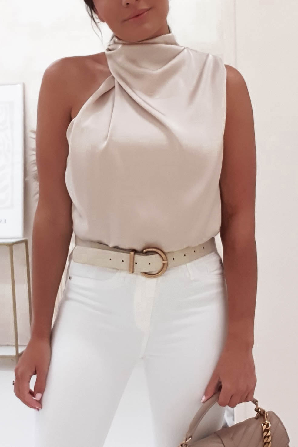 A woman in a sleeveless, high-neck beige top and white pants with an elasticated waist stands holding a tan handbag with a chain strap. She is wearing a beige belt with a gold buckle. The background is softly blurred.