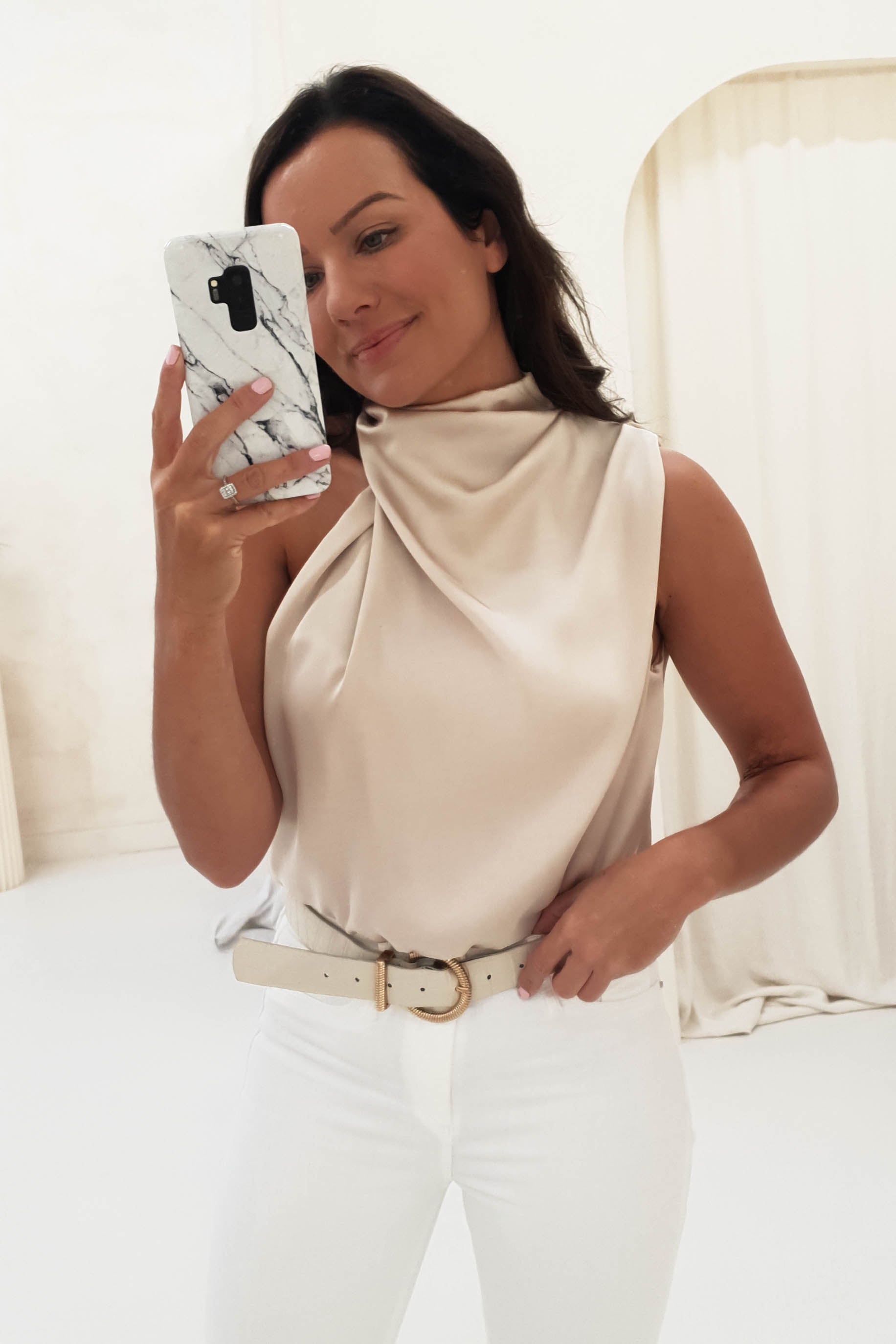 A woman is taking a mirror selfie in a minimalist room, wearing a beige, sleeveless, draped top and white pants with an elasticated waist. Her phone, adorned with a marble-patterned case, captures her soft smile.