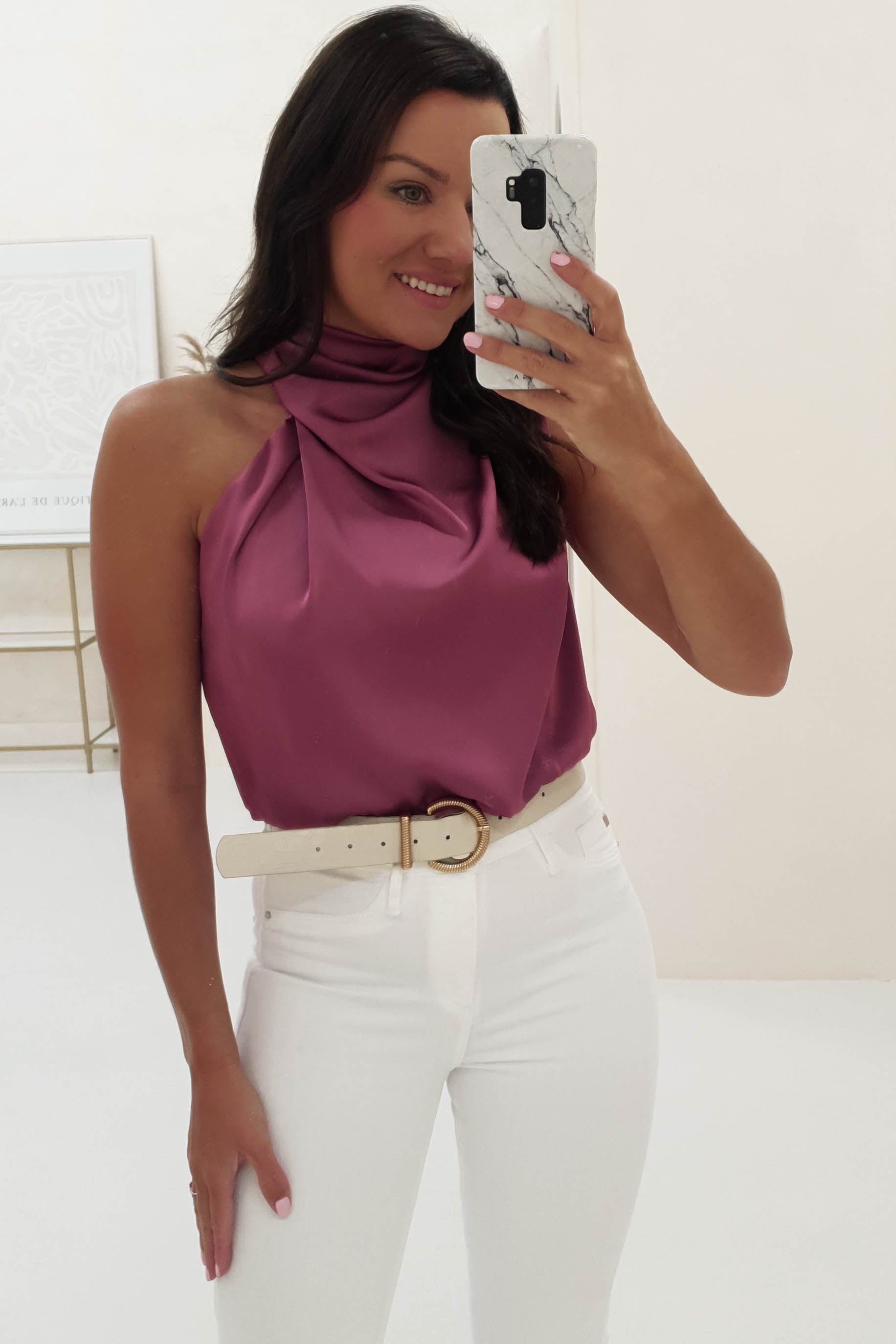 Woman taking a mirror selfie in a bright, minimalistic room, wearing a sleeveless mauve satin top with a draped neckline—crafted from luxurious satin fabric—paired with white high-waisted pants and a cream belt with a gold buckle.