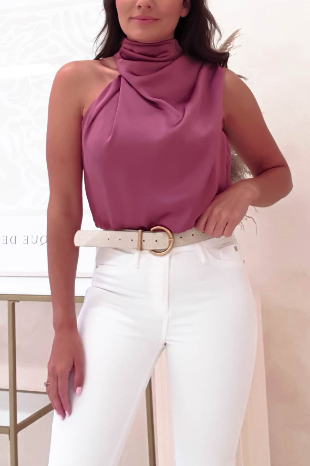 A woman wearing a sleeveless mauve halter top with a draped neckline, white high-waisted pants with an elasticated waist, and a white belt with a gold buckle, standing indoors against a neutral background.