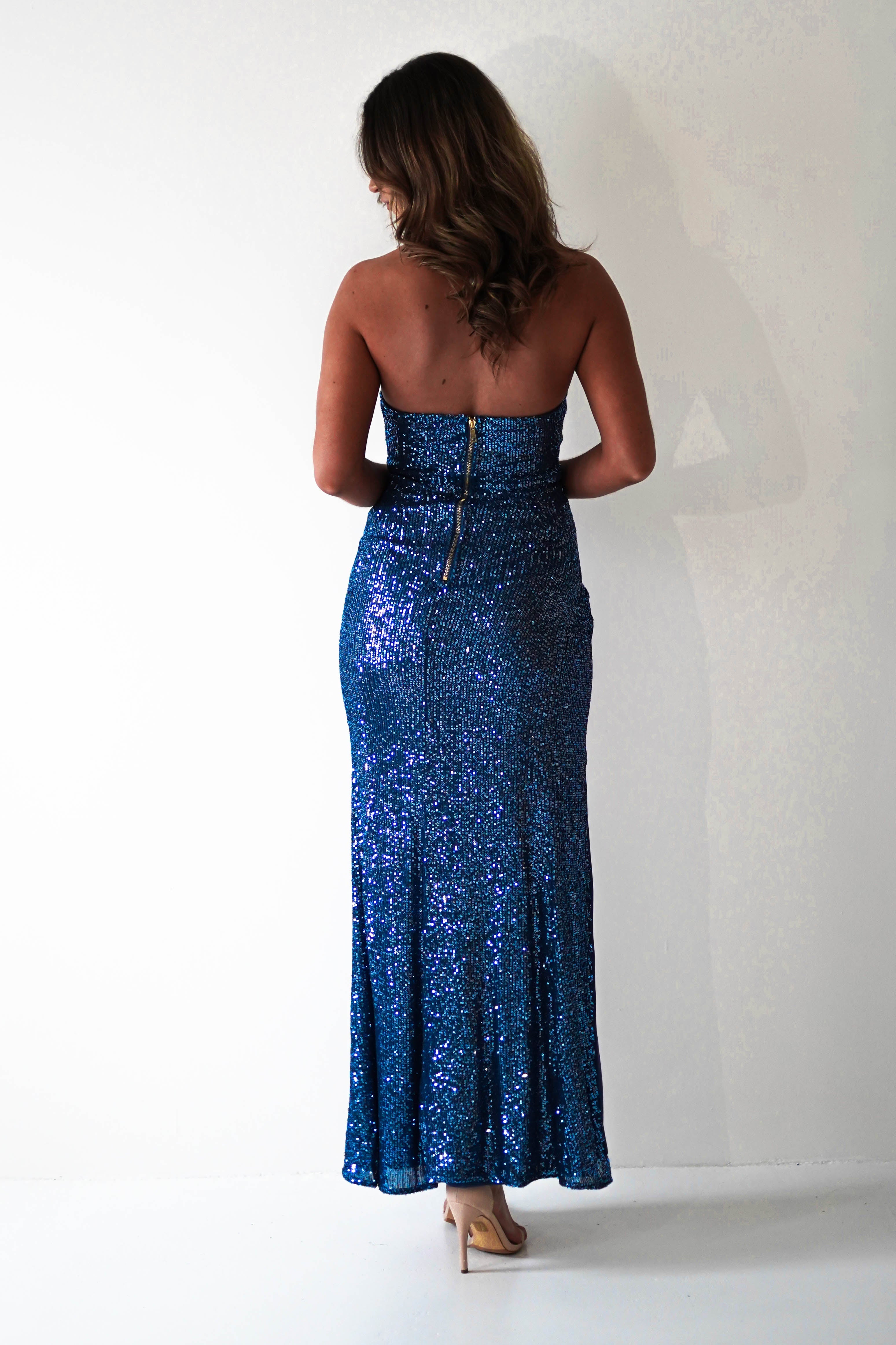 A woman stands facing away, wearing a strapless maxi dress with blue sequins and a visible back zipper—perfect for formal events. She has medium-length wavy brown hair and nude heels, standing against a plain white background.